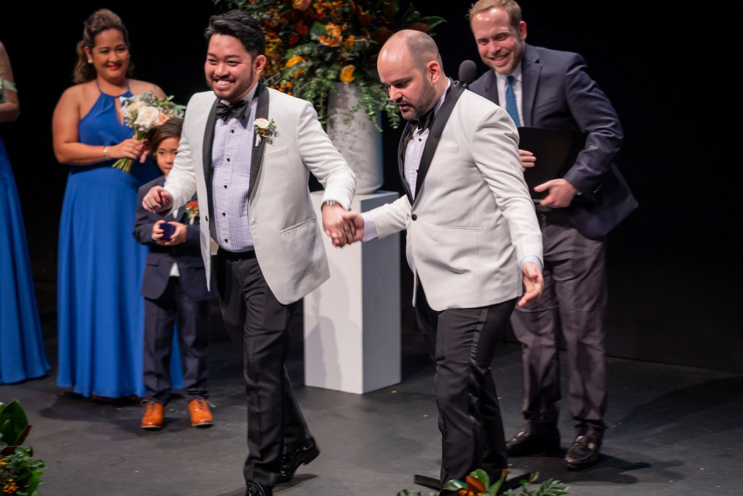 Two grooms in white tuxedo jackets share a joyful moment at their wedding ceremony with guests cheering behind them.