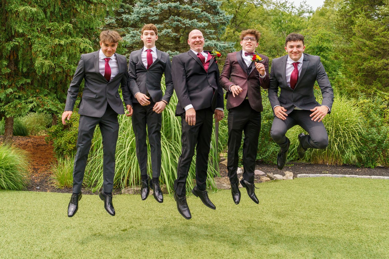Five groomsmen in dark suits and red ties jumping joyfully on a green lawn at an outdoor wedding celebration.