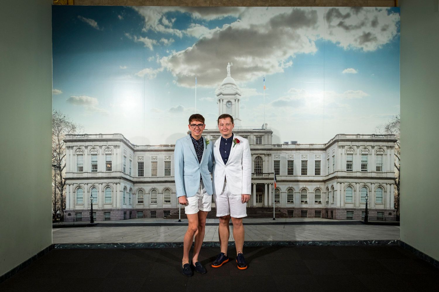 Two men in matching light blue blazers and white shorts pose in front of a grand building backdrop.