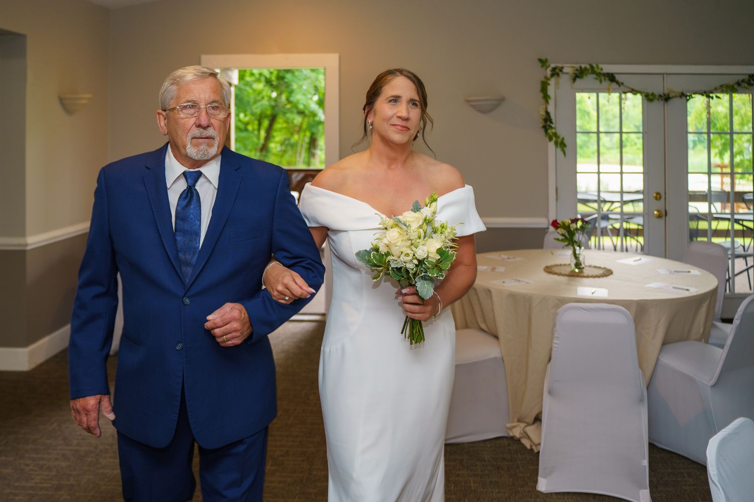 Bride in white off-shoulder gown walks down aisle with older man in blue suit, holding bouquet.
