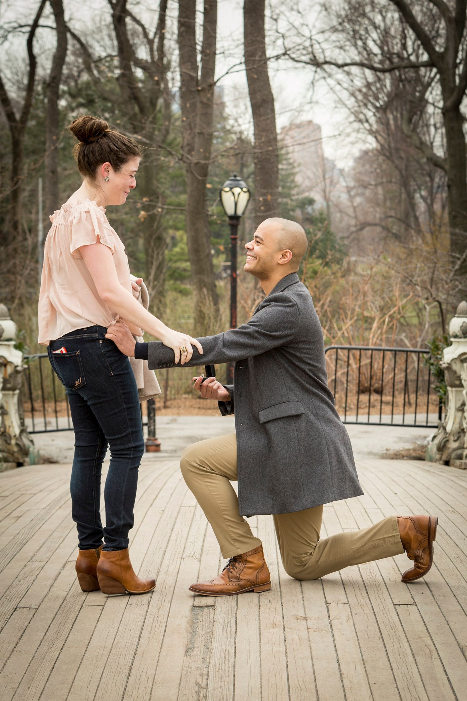 Man kneeling on one knee proposing to woman on wooden bridge in park, romantic marriage proposal outdoors.