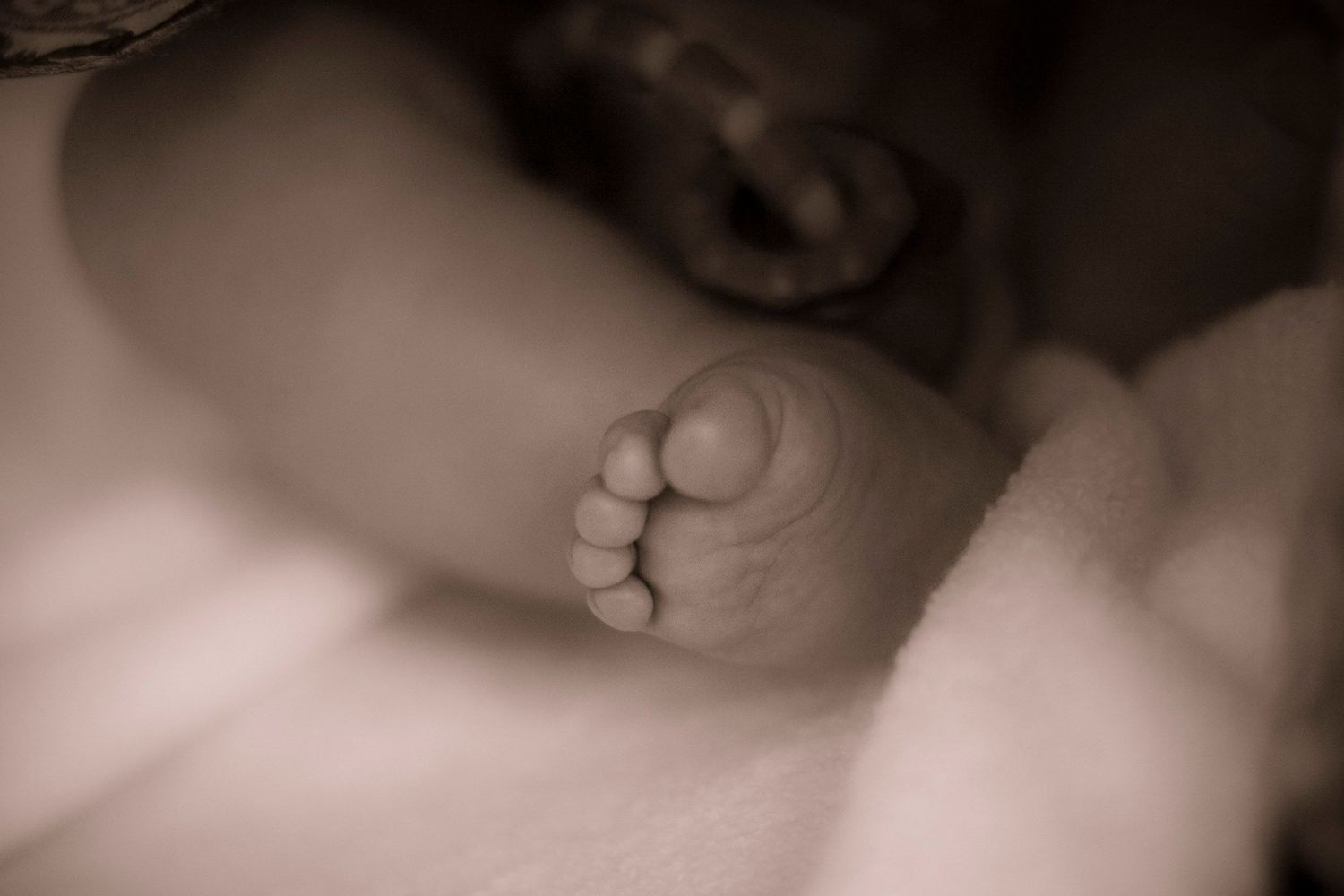 Close-up sepia photo of a newborn baby's tiny foot resting on a soft white blanket.