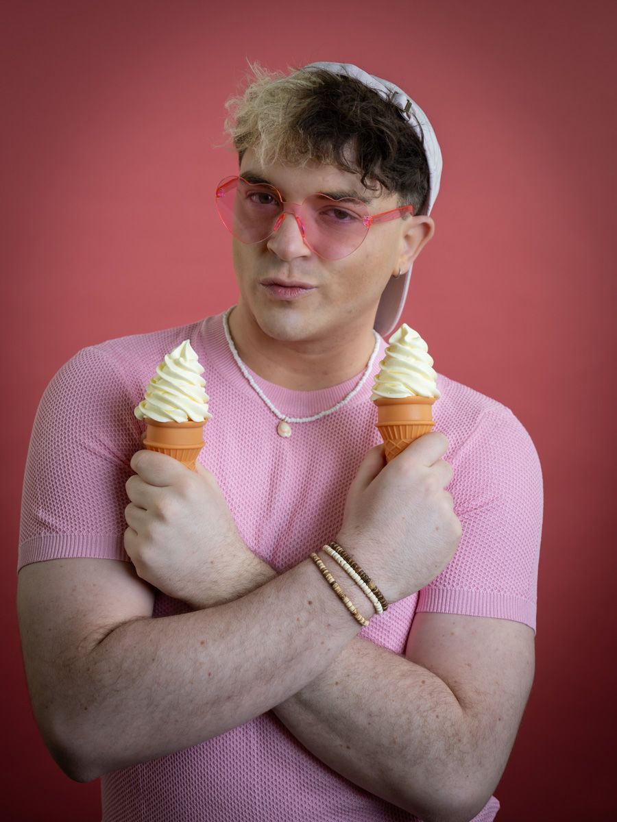 Man in pink mesh shirt and pink sunglasses holding two soft serve ice cream cones crossed over his chest against pink background.
