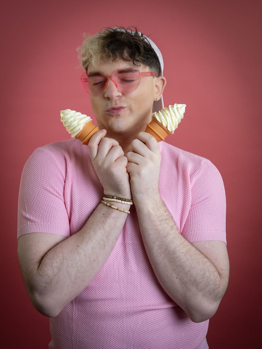 A young man in a pink shirt and pink sunglasses holds two ice cream cones against a red background.