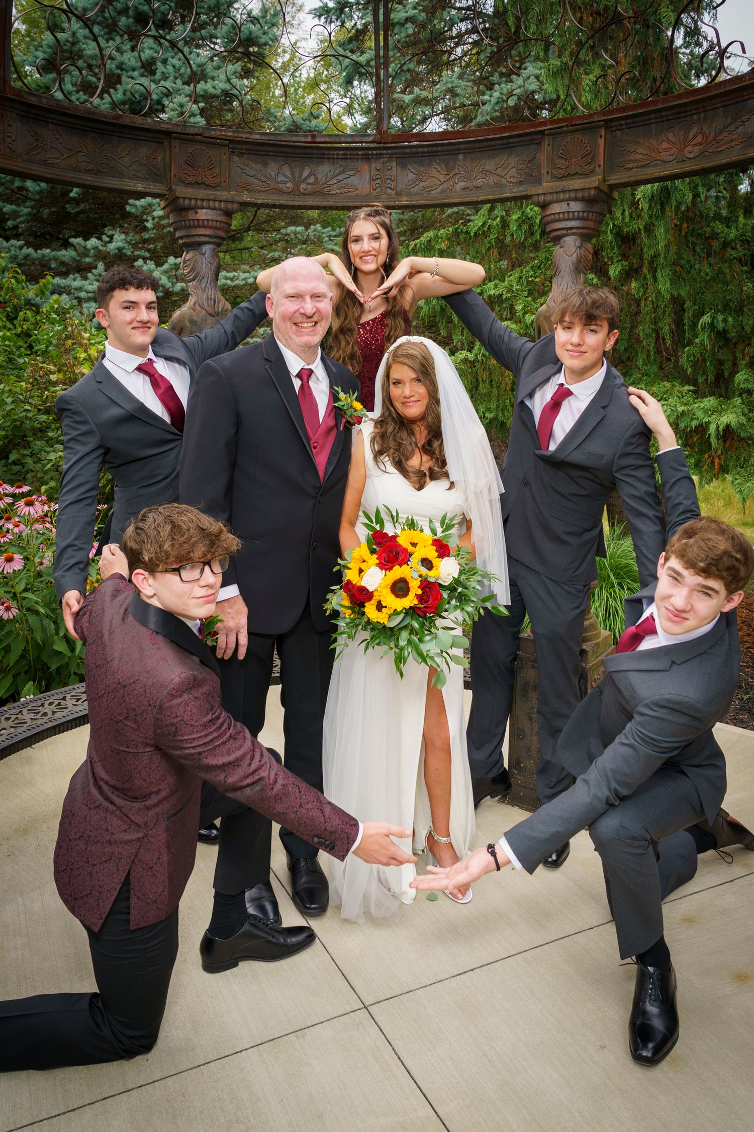 Wedding couple poses with groomsmen and bridesmaid in a garden gazebo, bride holding sunflower bouquet.