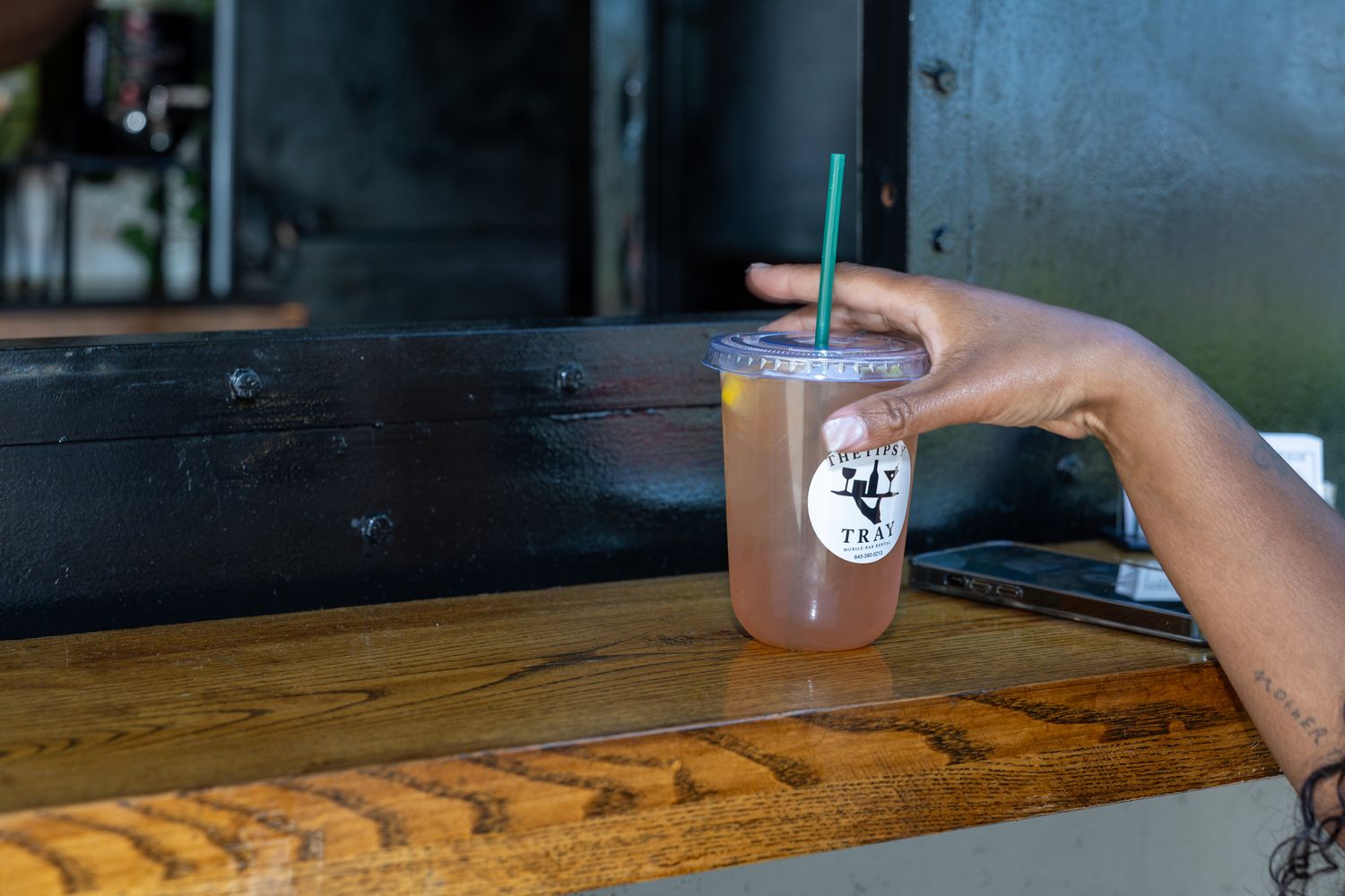 A hand holds a pink lemonade drink with a green straw at an outdoor food vendor window.