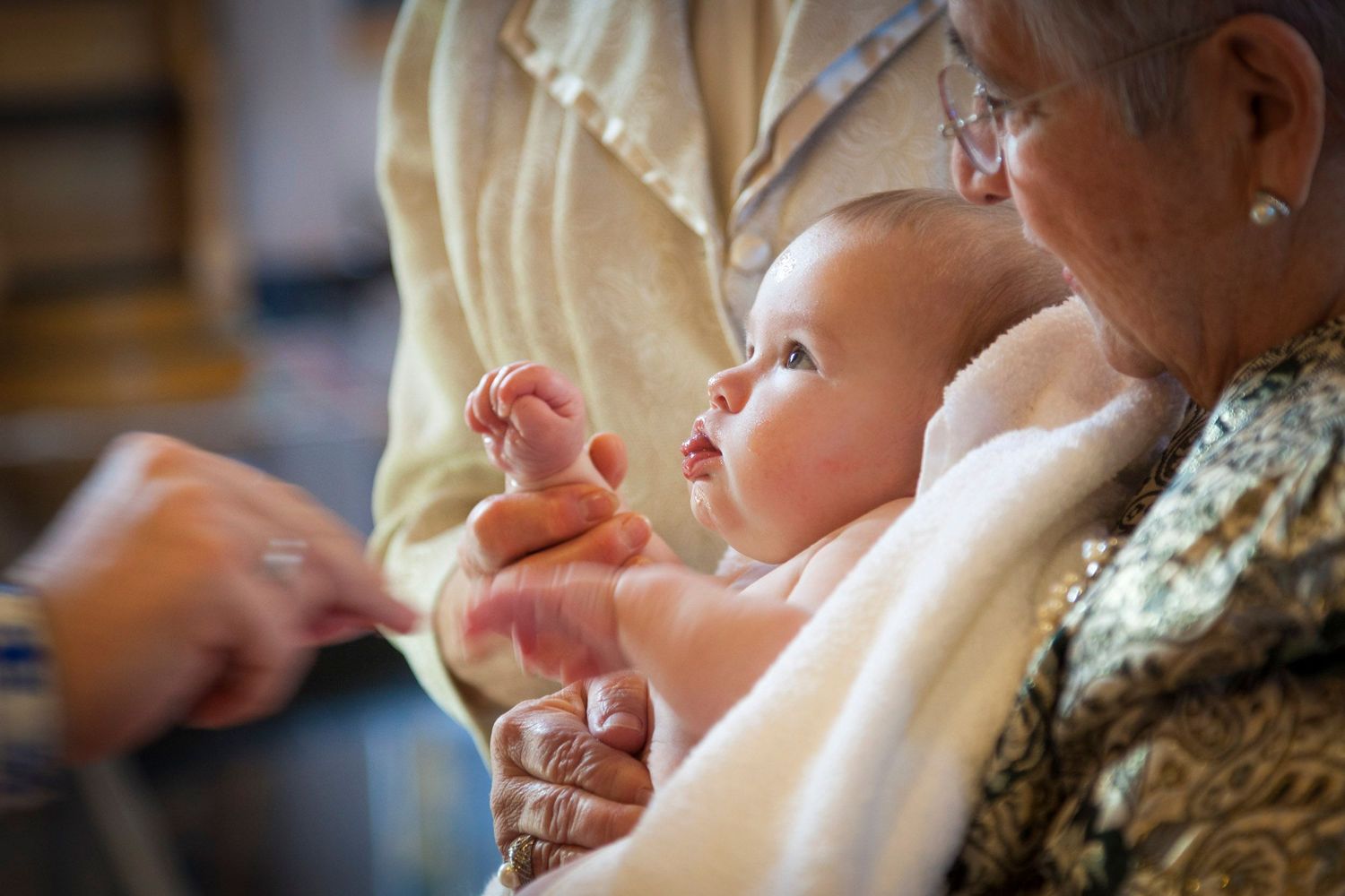 Elderly woman holding a newborn baby wrapped in a white blanket during a christening ceremony.