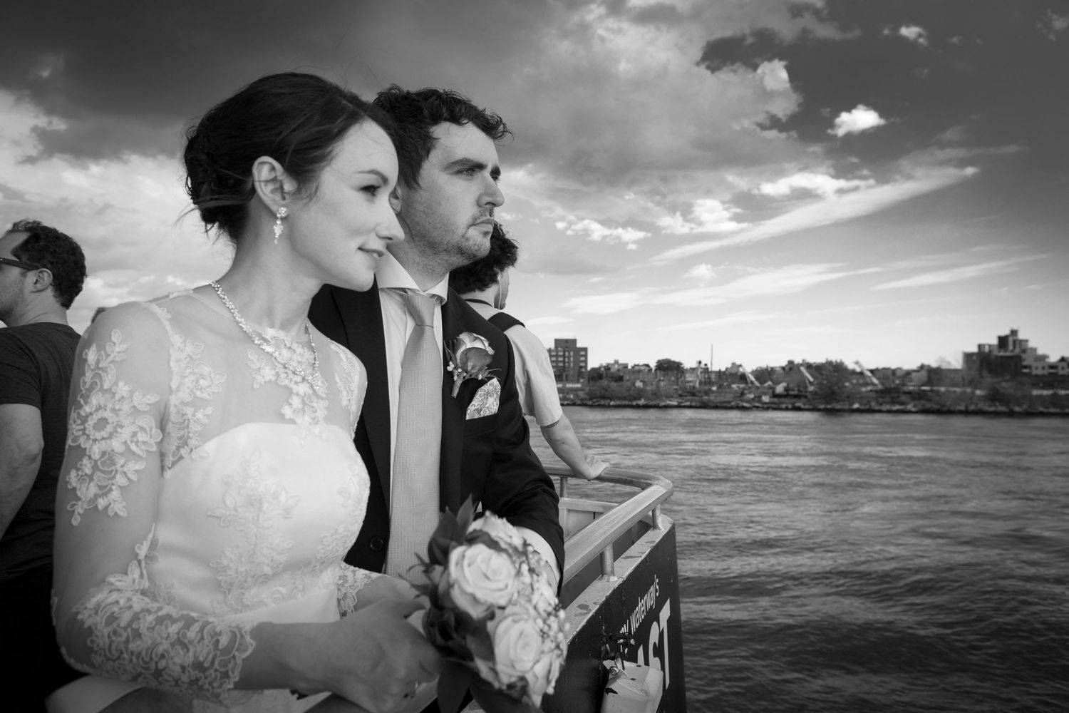 Black and white photo of a bride and groom standing by a river on their wedding day, bride holding a bouquet.