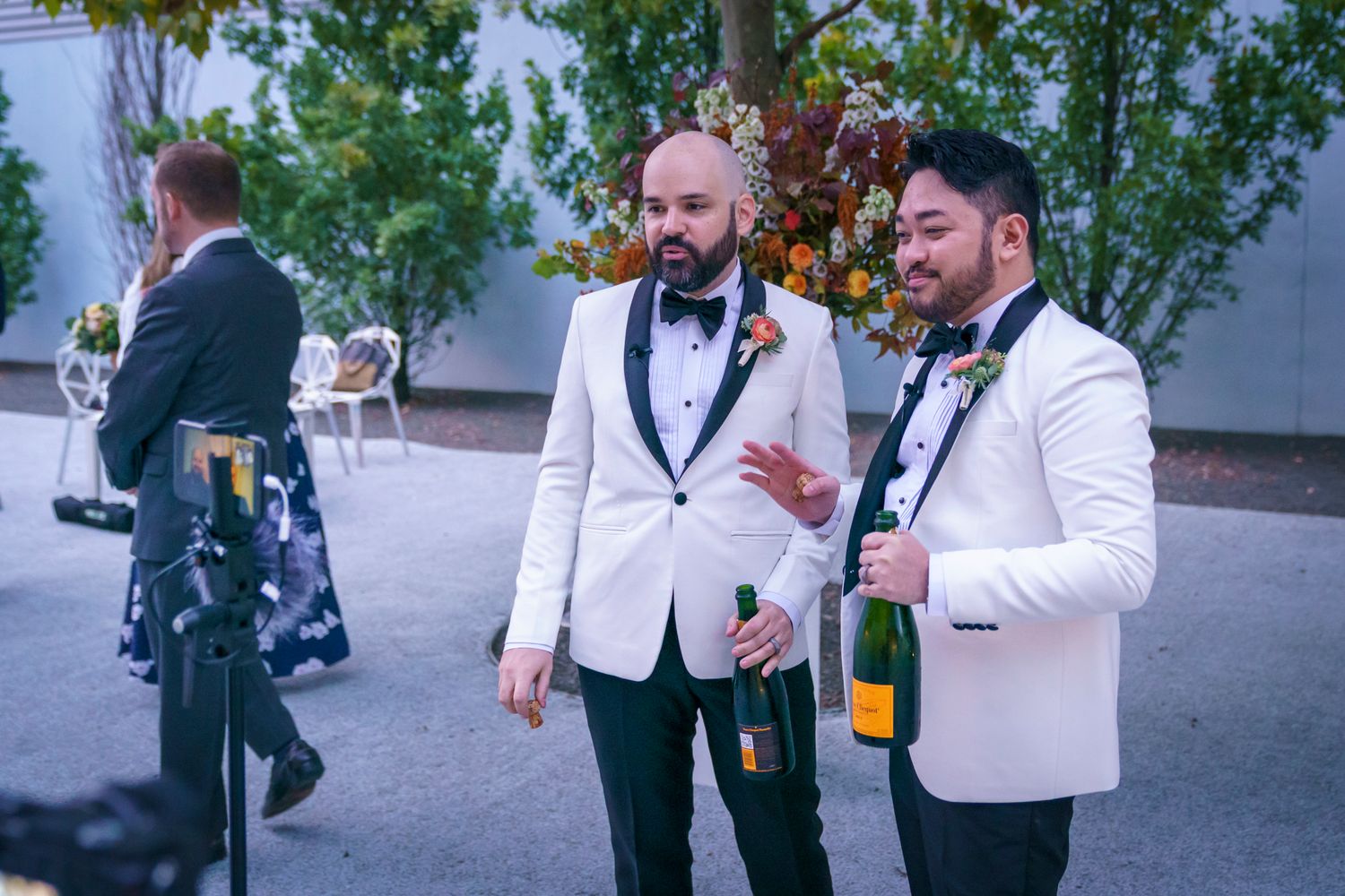 Two grooms in white tuxedos celebrate outdoors holding champagne bottles at their wedding reception.