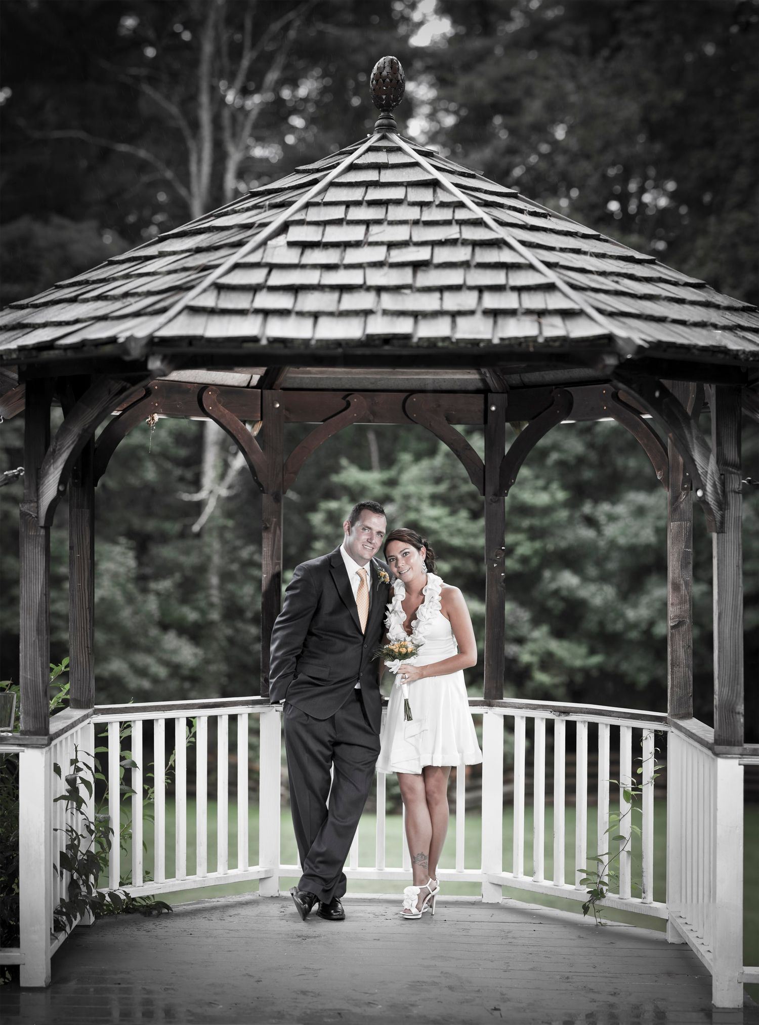 Wedding couple posing romantically inside white gazebo surrounded by lush green trees.