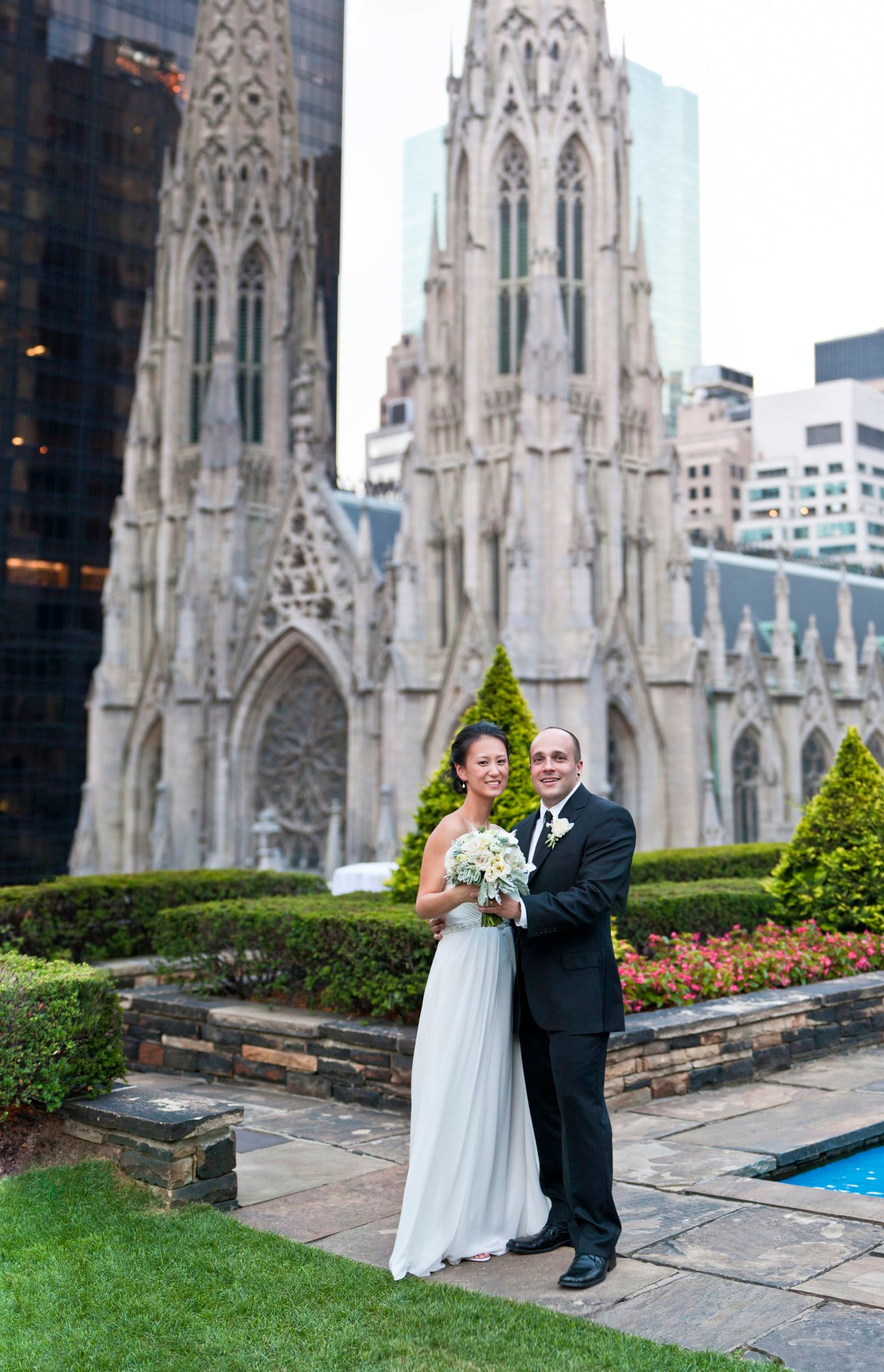 Bride and groom pose together on rooftop garden with Gothic cathedral architecture in background.