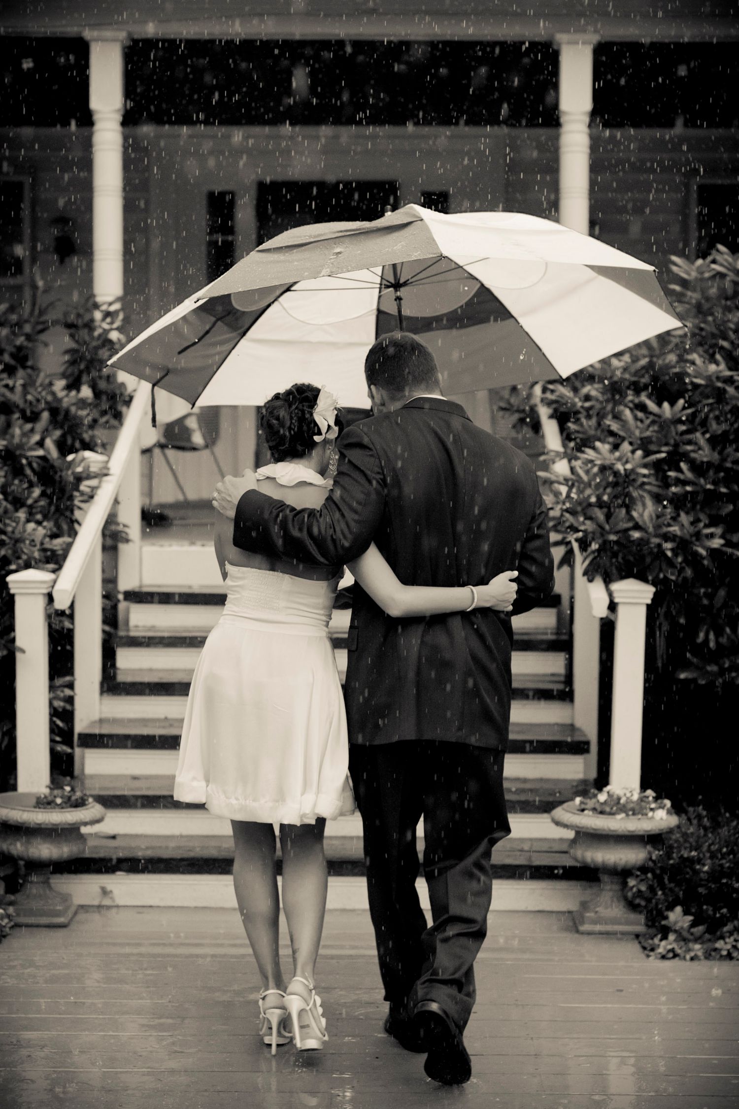 Couple sharing an umbrella in the rain walking toward a house in black and white.