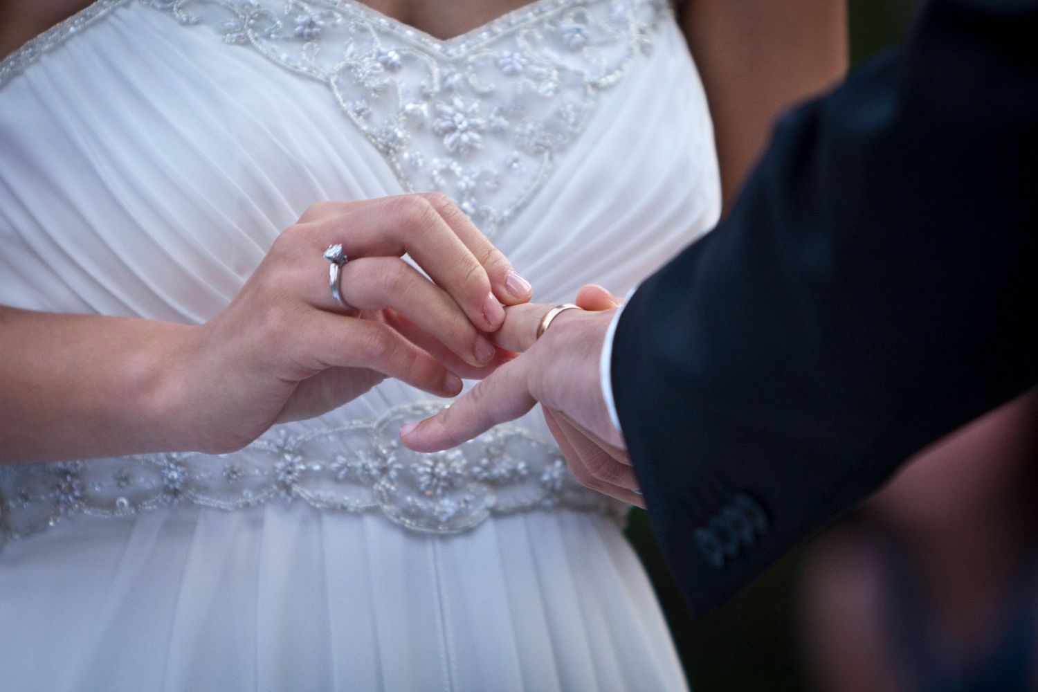 Bride in lace wedding dress places gold ring on grooms finger during wedding ring exchange ceremony.