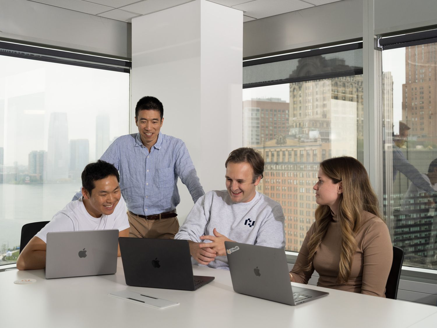 Four professionals collaborate around laptops in a modern office with city skyline views.