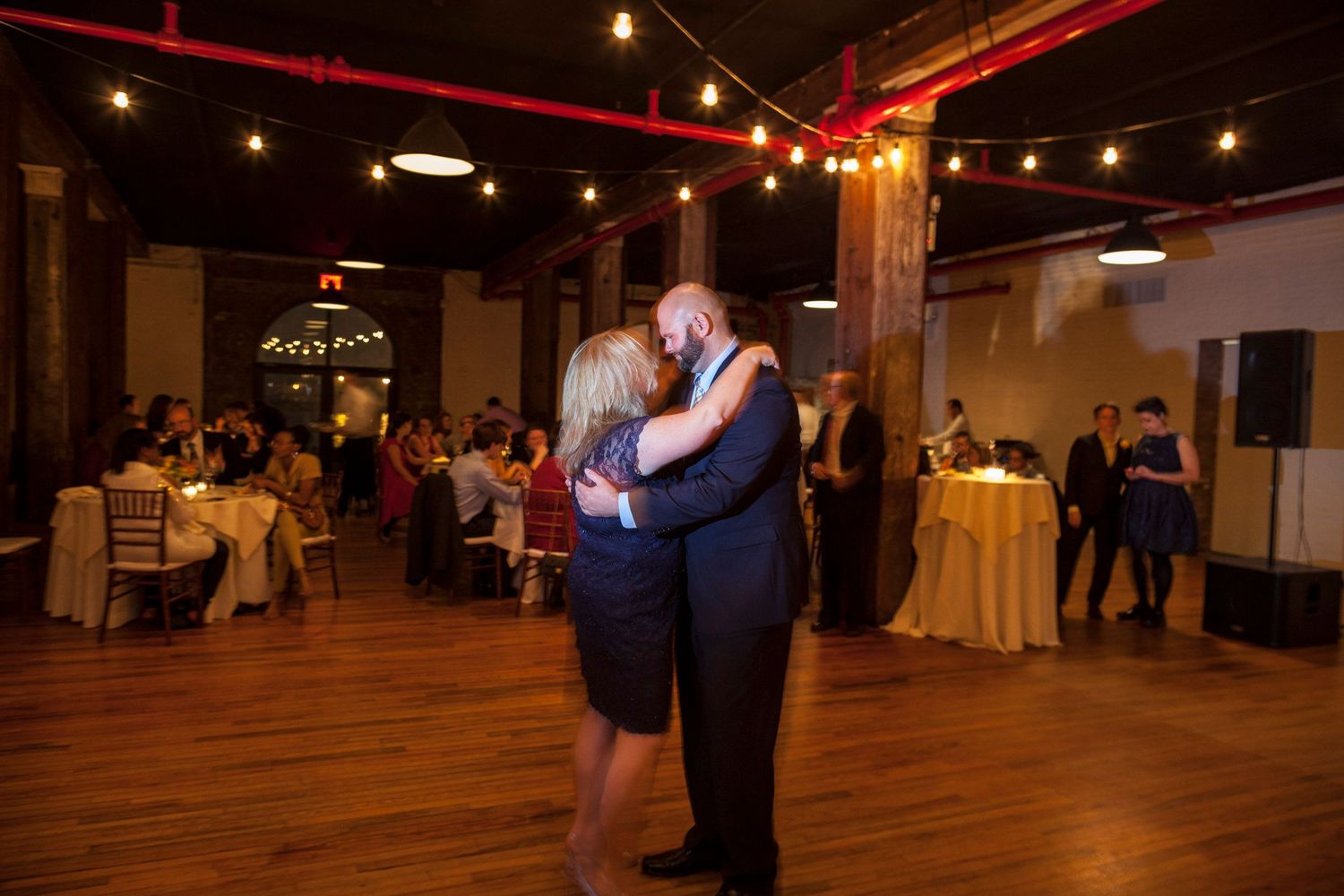 Couple shares romantic first dance at elegant wedding reception venue with string lights and guests seated at tables.