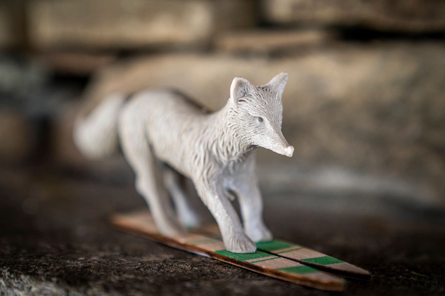 White fox figurine standing on a small wooden platform with green detail, displayed on a rustic stone surface.
