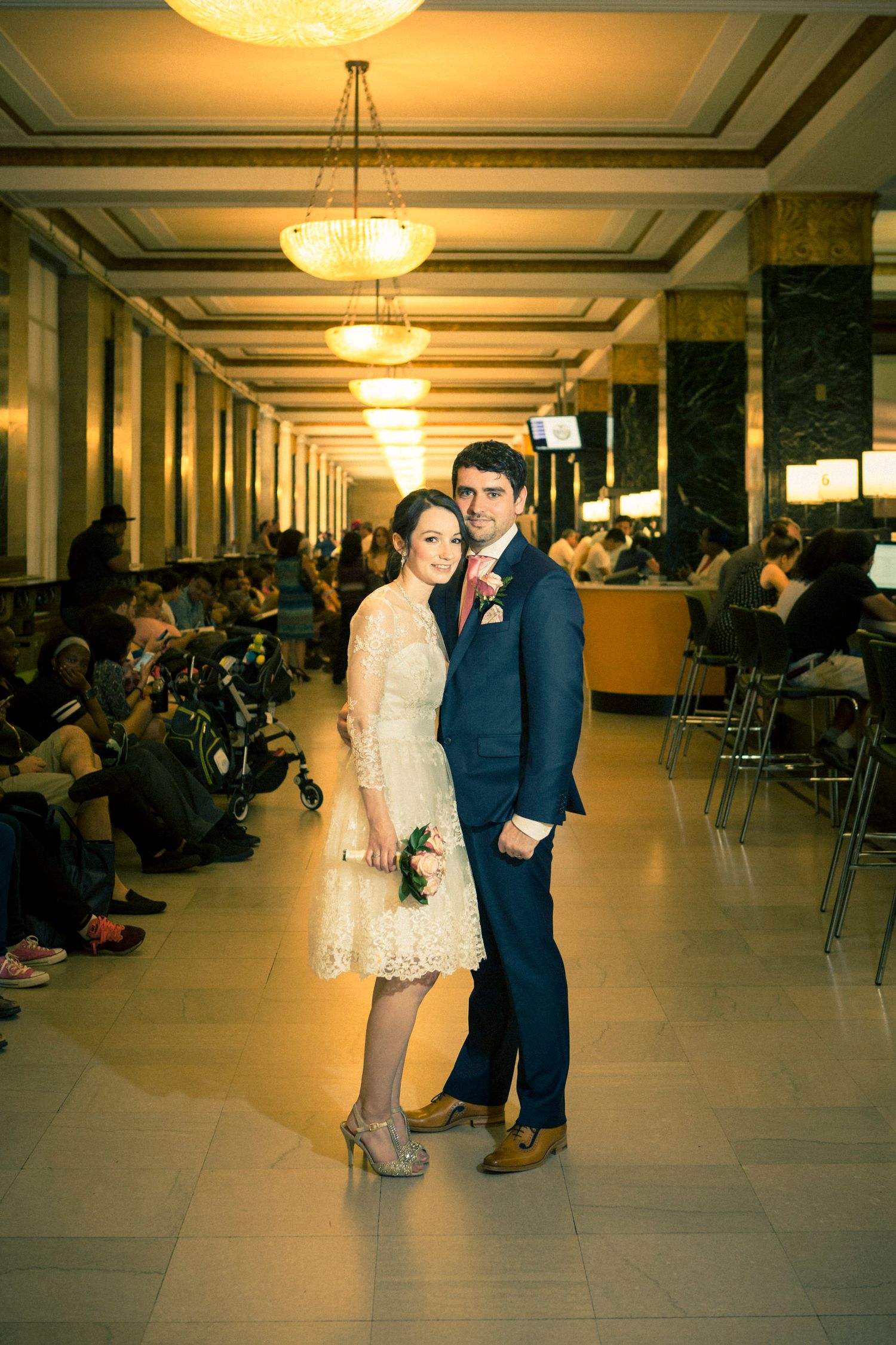 Bride and groom posing in elegant wedding attire inside a grand hall with ornate chandeliers.