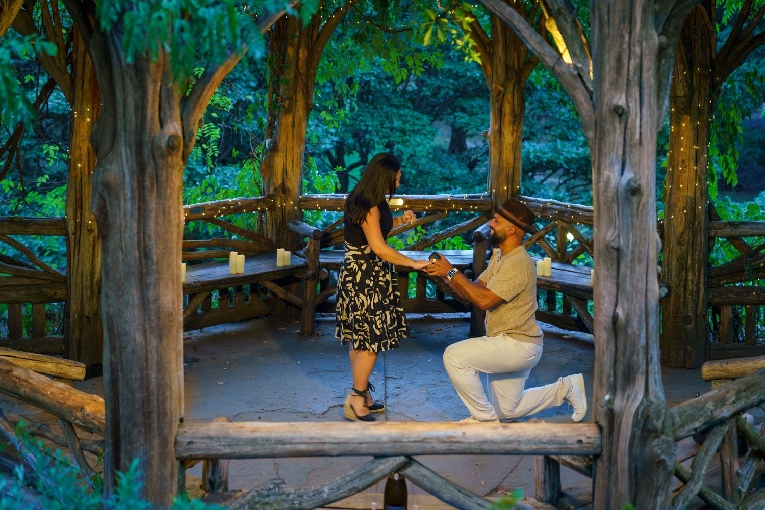 Man proposes to woman on one knee in a romantic rustic wooden gazebo surrounded by lush greenery and candlelight.