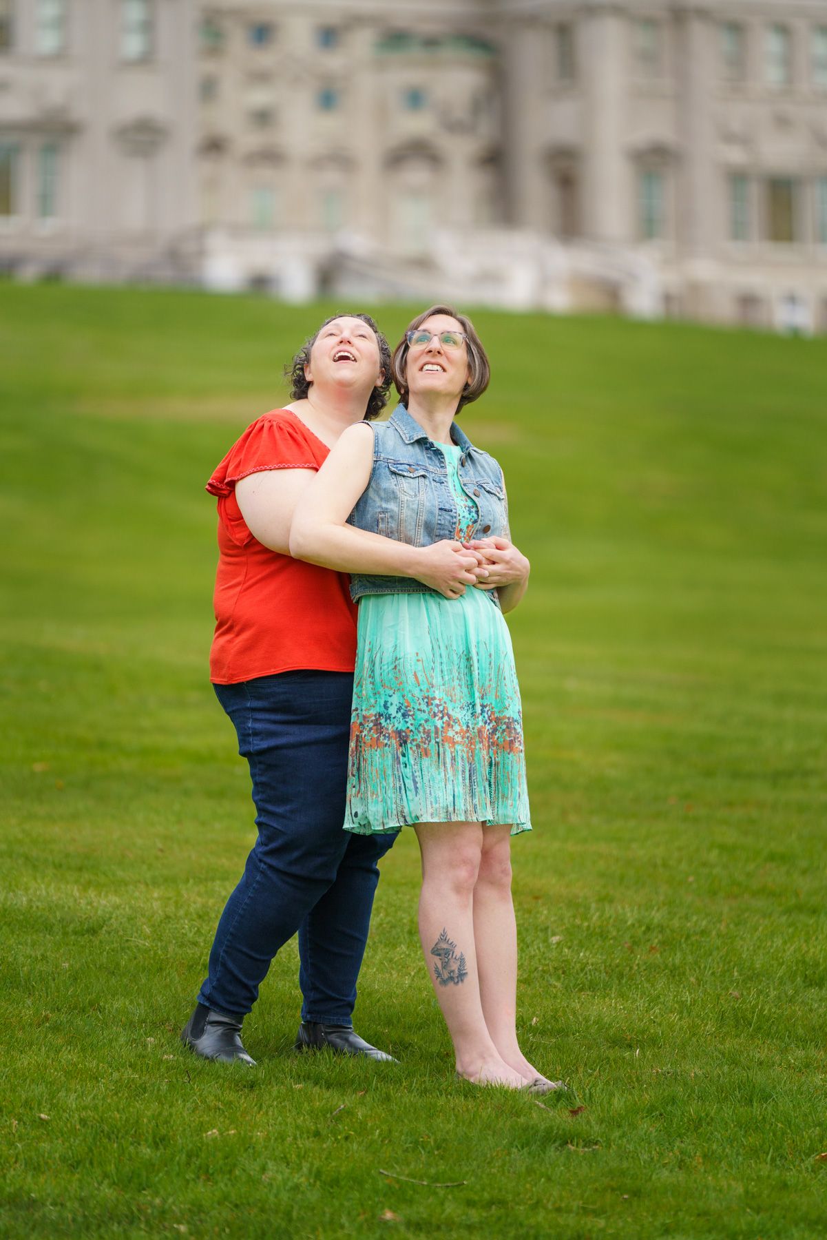 Two women embracing on green lawn, looking up joyfully, wearing red top and teal dress, with historic building behind.