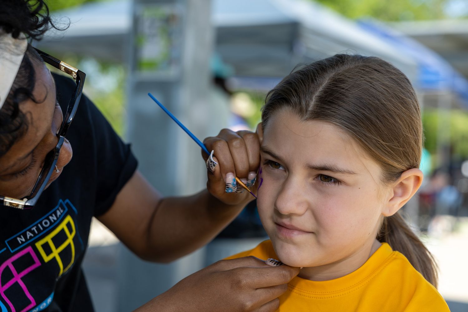 Artist applying face paint to a young girl in a yellow shirt at an outdoor event.