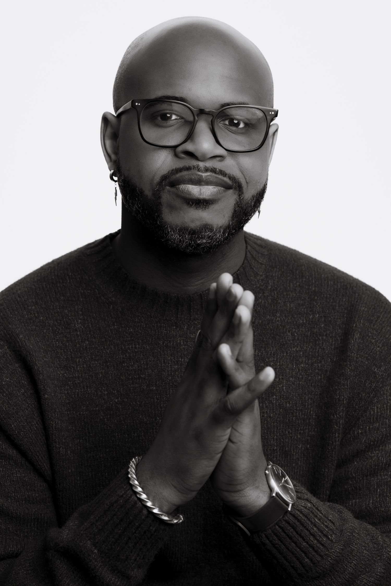 Black and white portrait of a bald man with glasses and beard, wearing a sweater, hands clasped together.