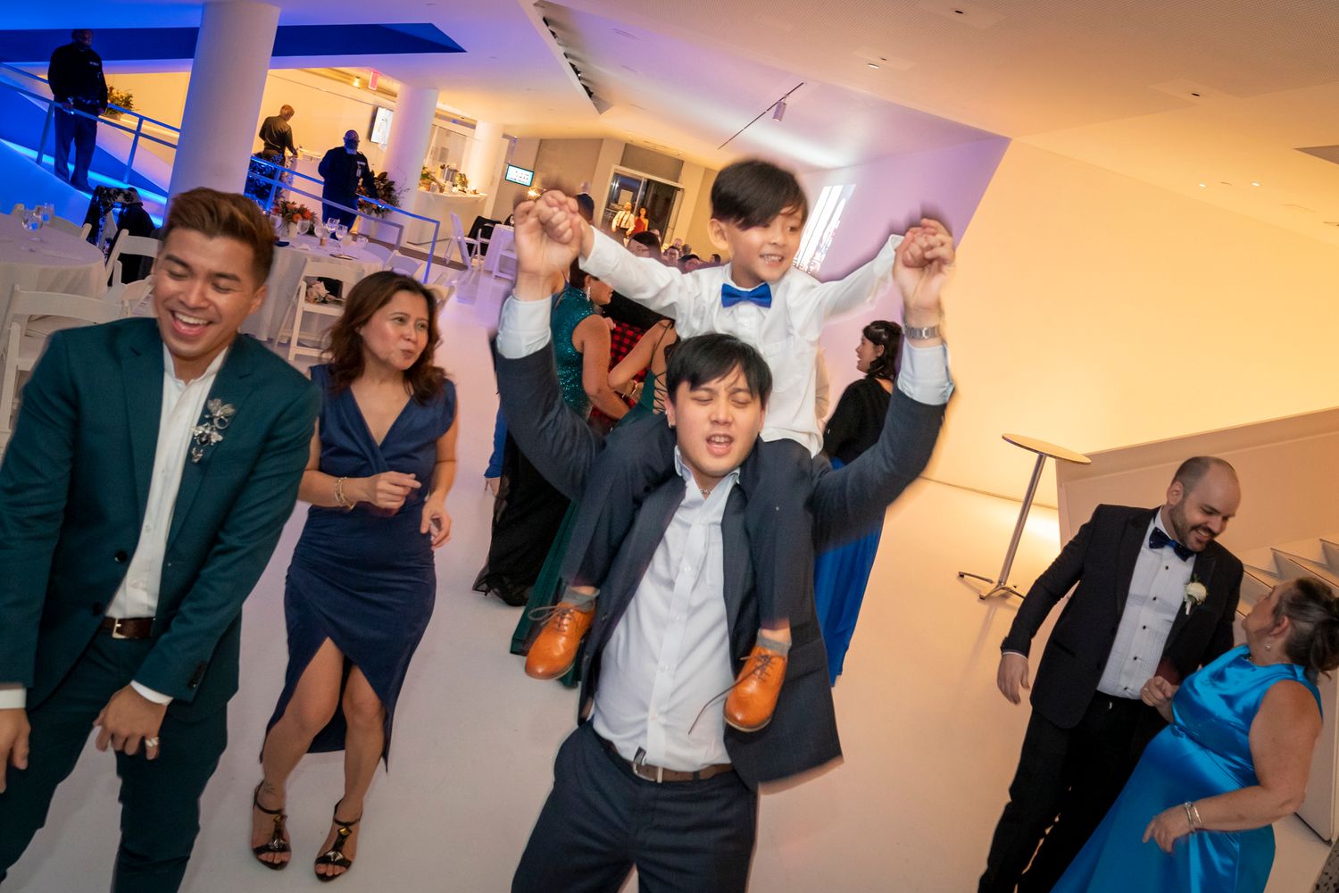 A joyful wedding reception scene with a child on a man's shoulders dancing on a white dance floor.