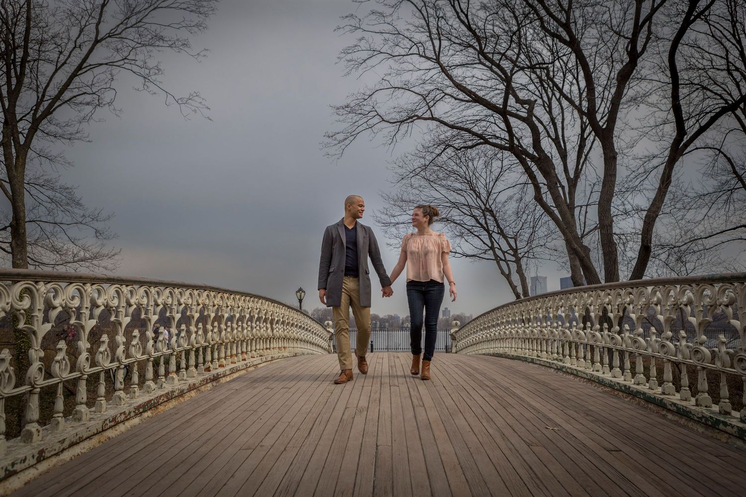 Couple holding hands walking on ornate bridge with bare trees and cloudy sky backdrop.