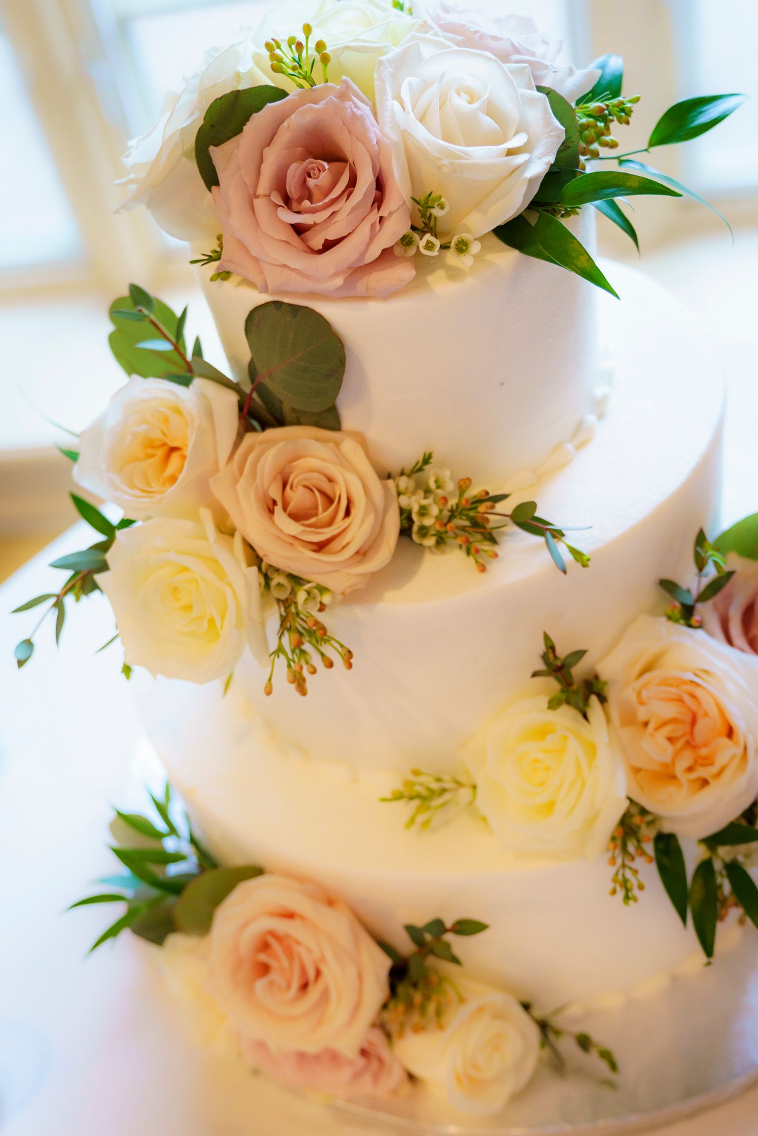 Elegant three-tier white wedding cake decorated with blush, cream, and yellow roses with greenery accents.