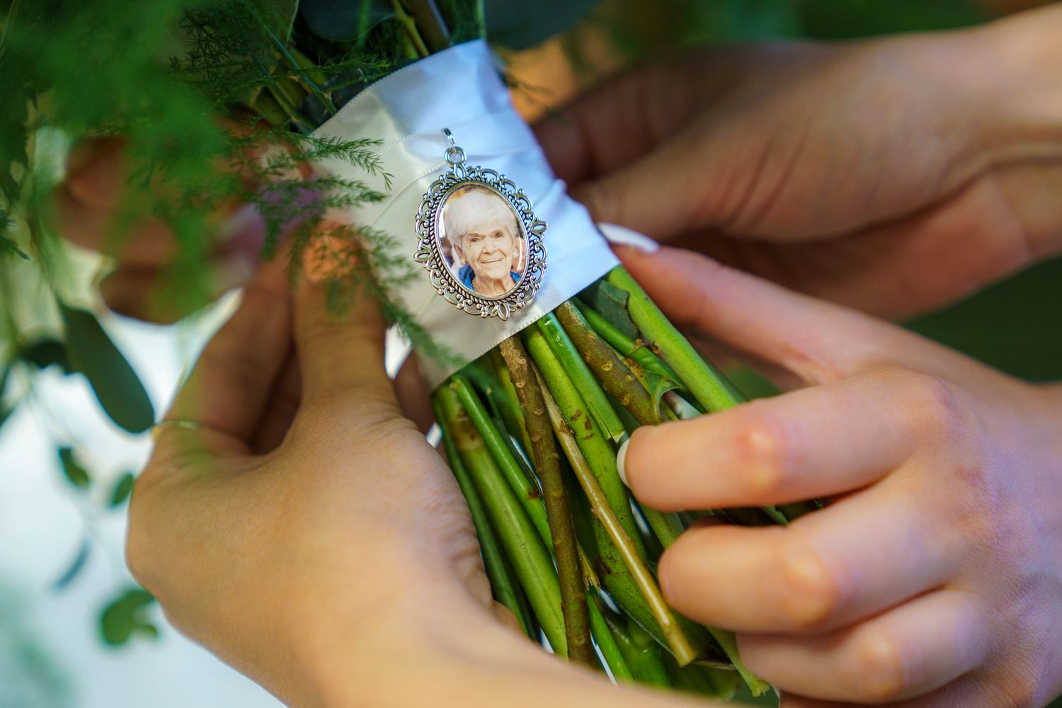Hands holding a floral bouquet with a memorial photo charm pendant attached to green stems.