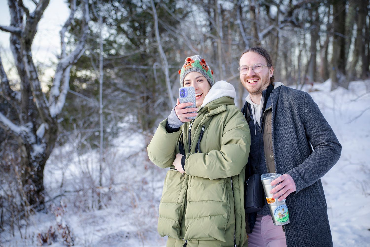 Couple smiling outdoors in snowy winter forest, woman in green parka holding phone, man in gray coat holding drink.