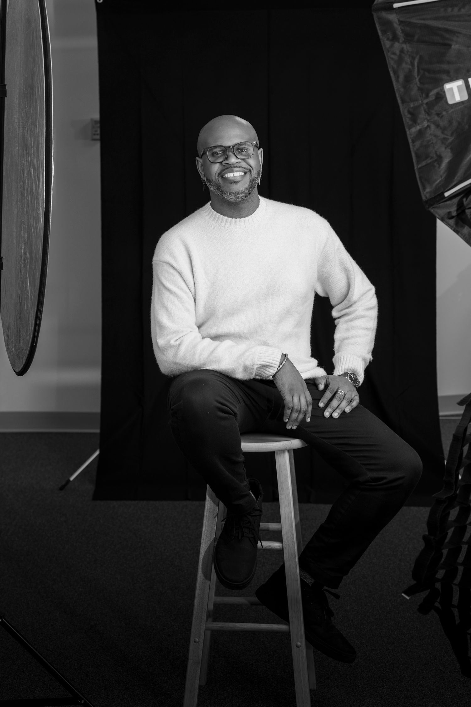Smiling bald man wearing glasses and white sweater sits on stool in professional black and white studio portrait.