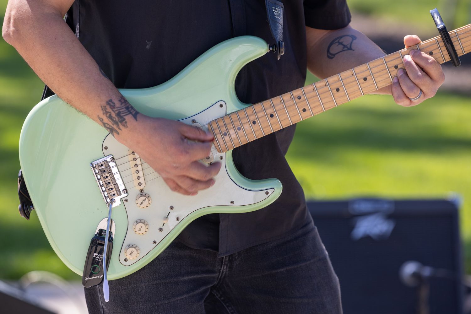 Tattooed musician playing a mint green Fender Stratocaster electric guitar outdoors at a live music event.