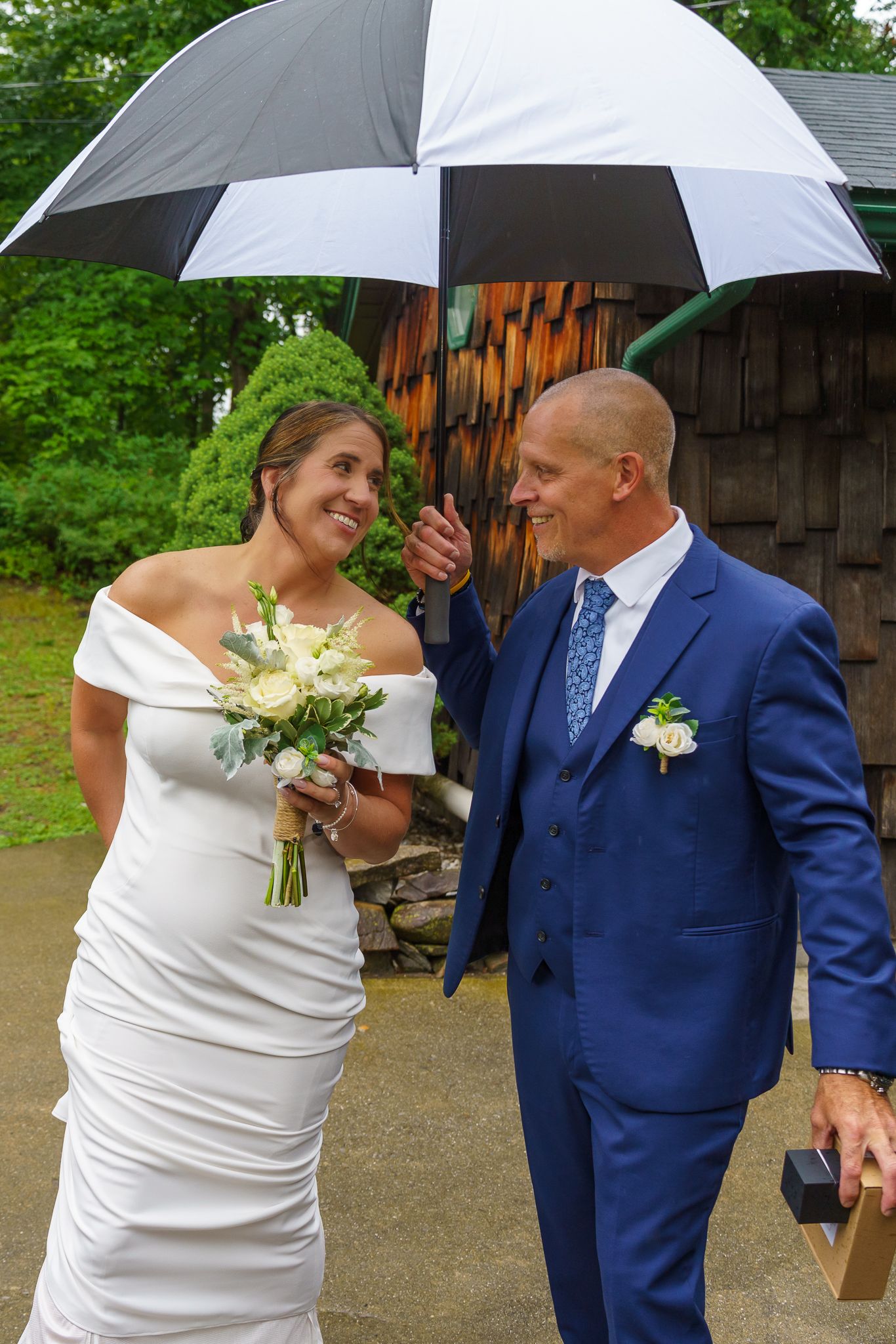 Bride in white gown holds bouquet while groom in blue suit holds black-and-white umbrella on rainy wedding day.