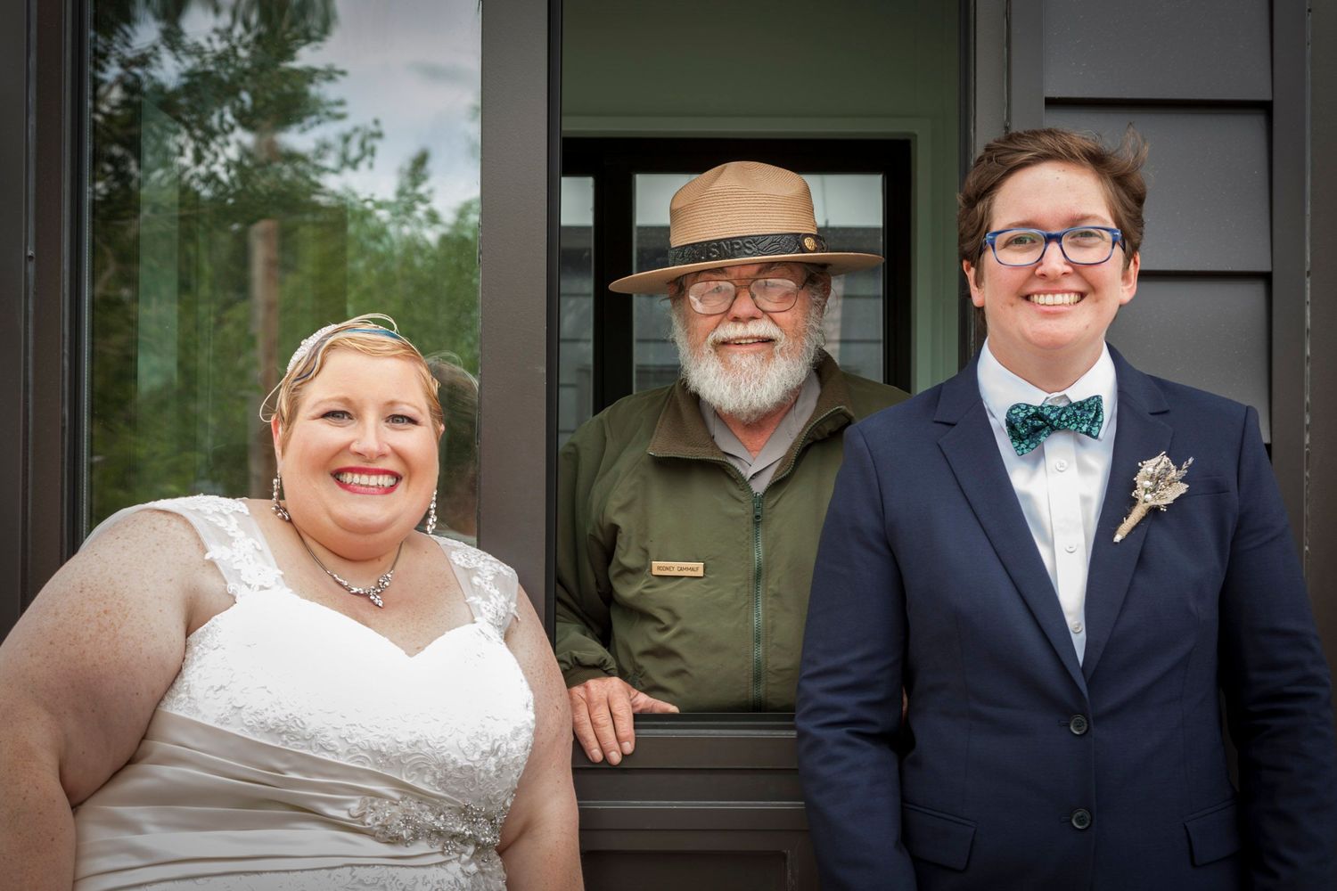 Two brides smile with a park ranger in uniform between them outside a wooden building on their wedding day.