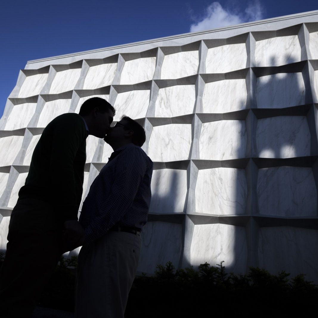 Silhouette of a couple kissing in front of a white geometric building facade against a blue sky.