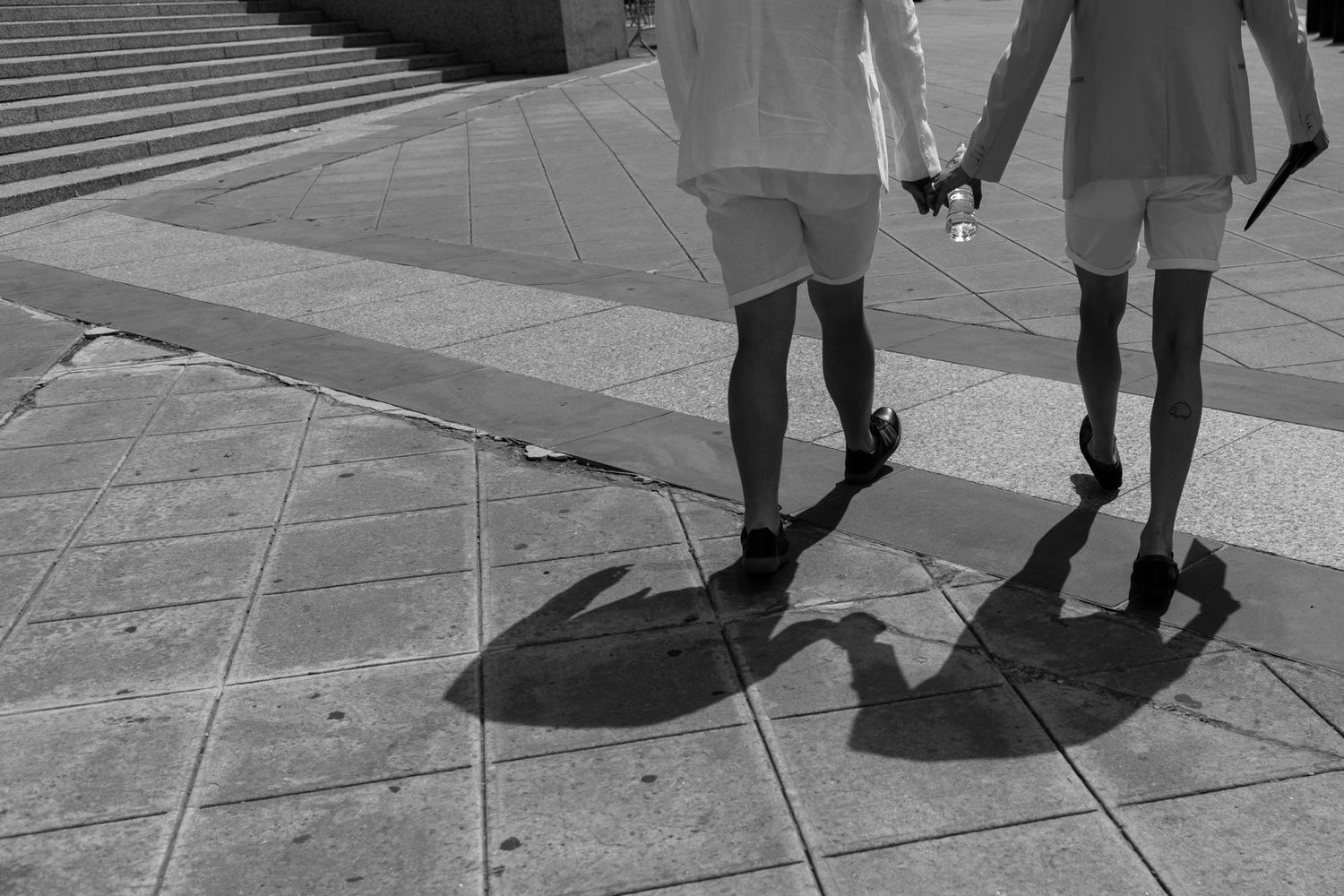 Black and white photo of two people holding hands, walking on a tiled plaza, casting long shadows in sunlight.