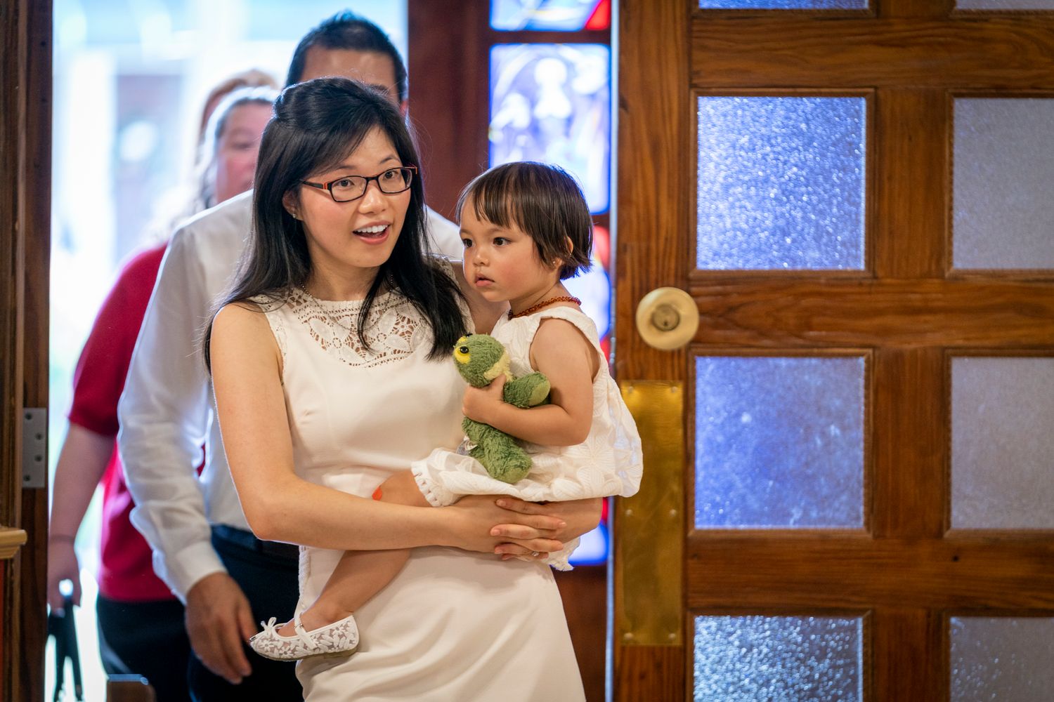 Woman in white dress holding toddler with stuffed animal at elegant venue entrance.