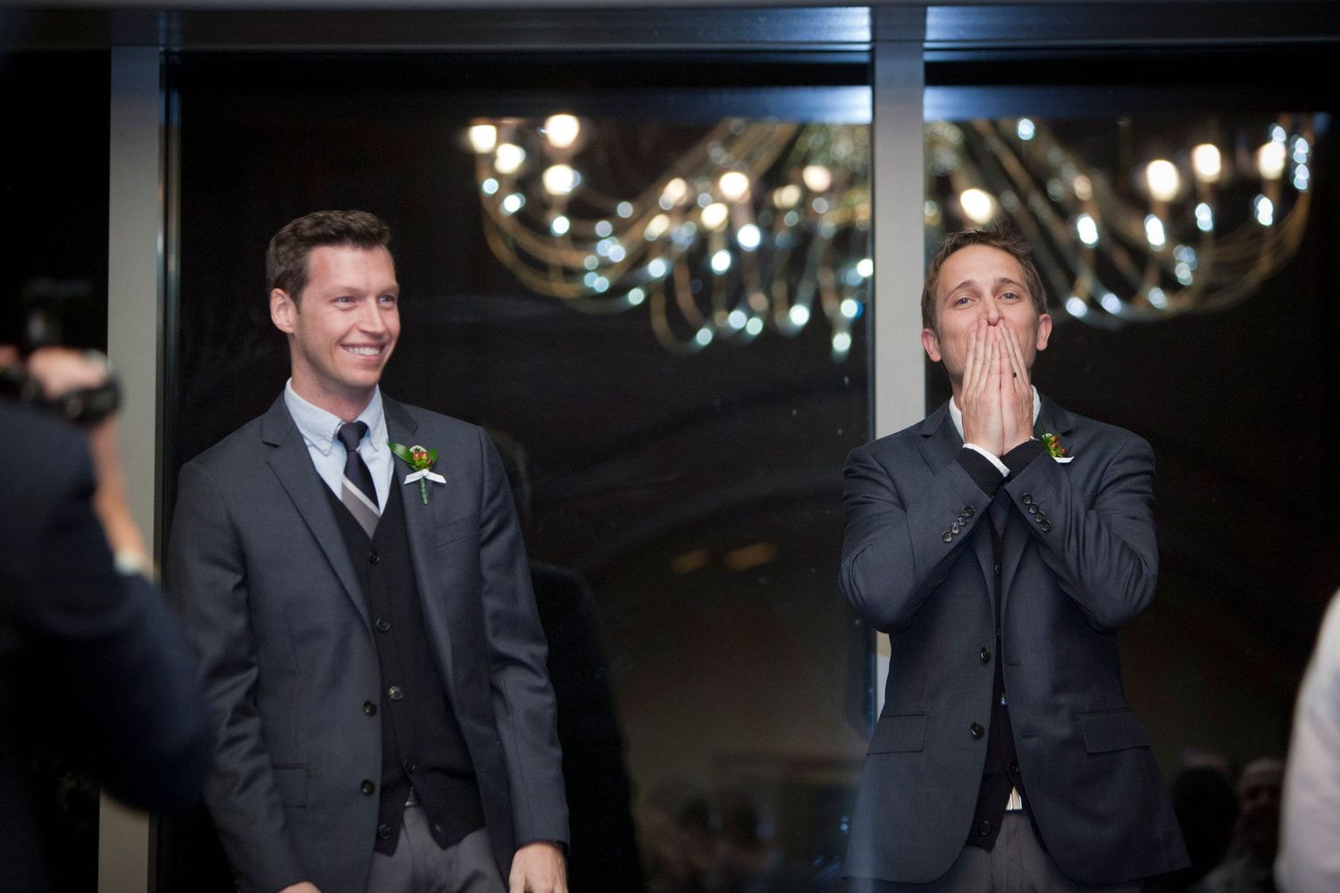 Two grooms in gray suits with yellow boutonnieres sharing a joyful moment at wedding.