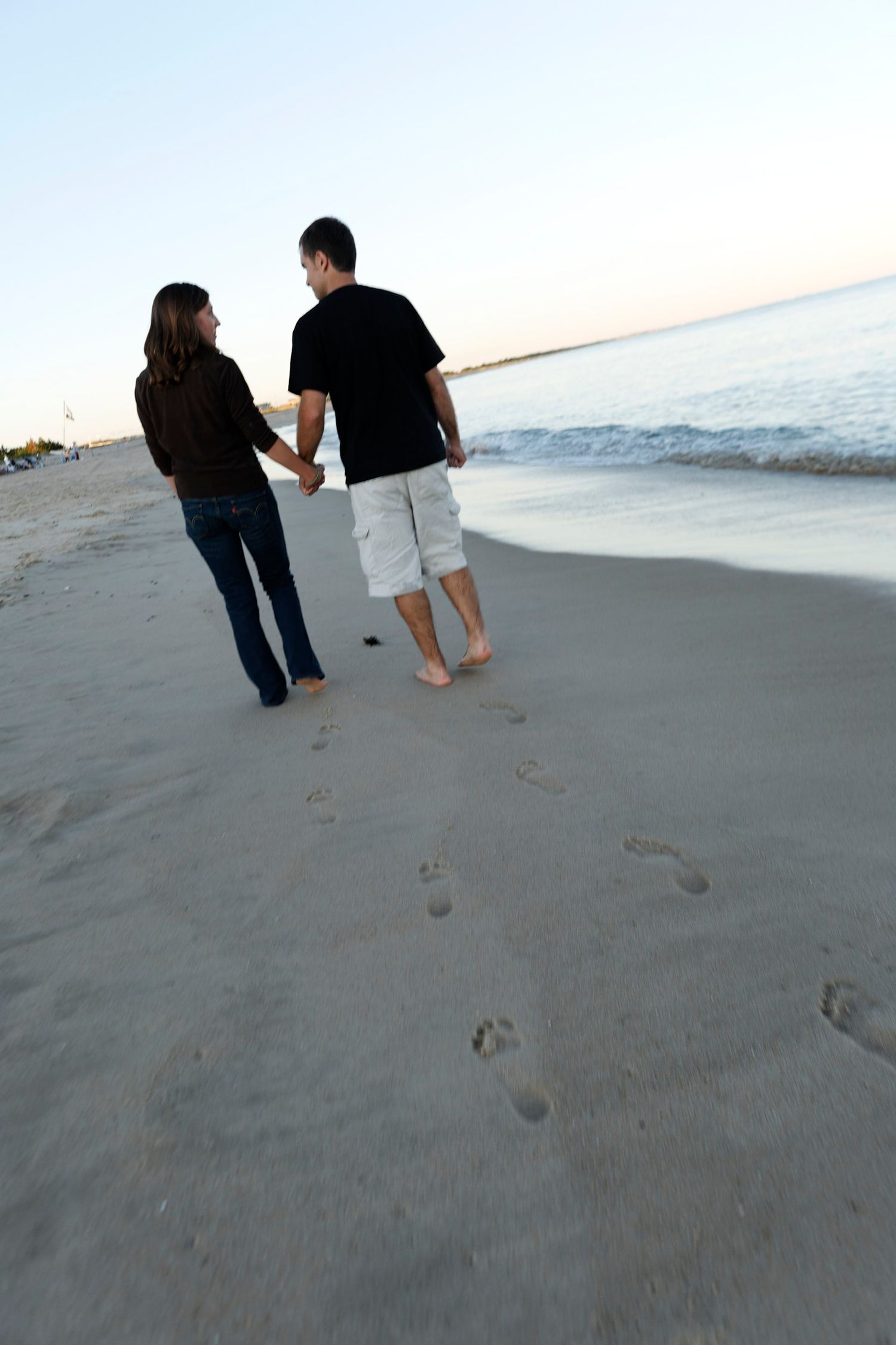 Couple holding hands walking barefoot on sandy beach leaving footprints at dusk.