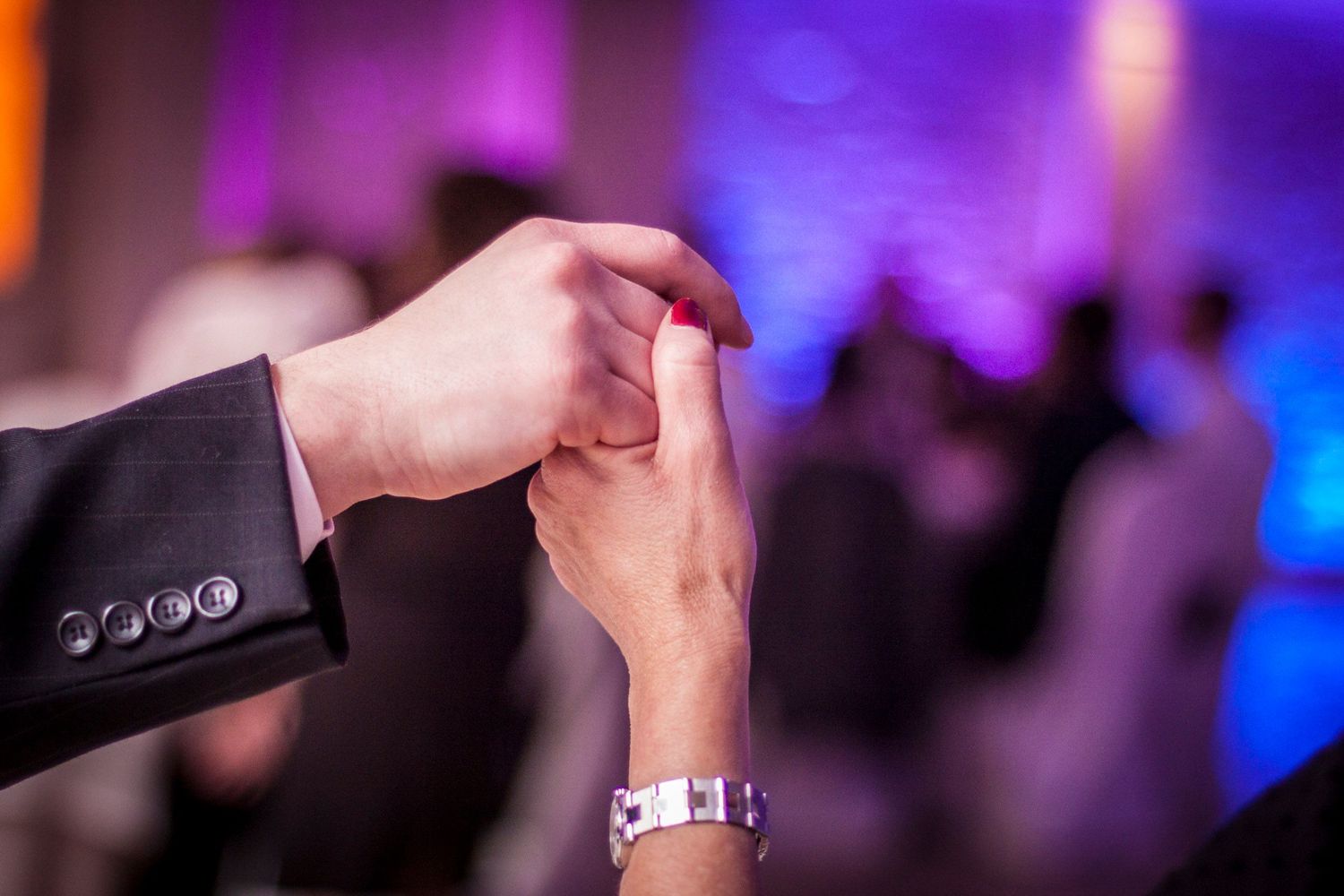 Couple holding hands on dance floor at wedding reception with colorful purple lighting.