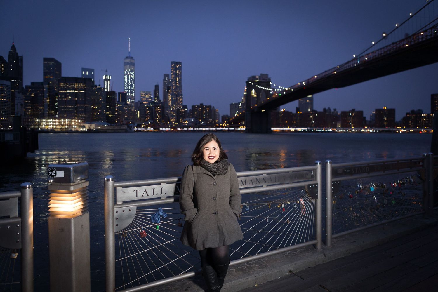 Woman in gray coat standing at Brooklyn waterfront with NYC skyline at night.