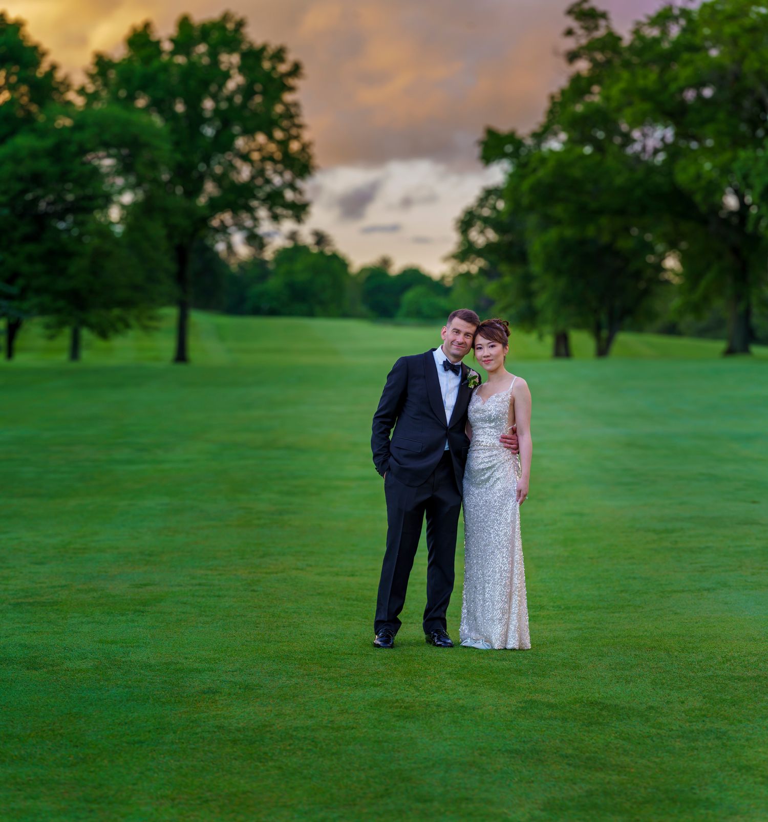 Bride and groom in formal attire standing on lush green golf course fairway beneath a stunning colorful sunset sky.