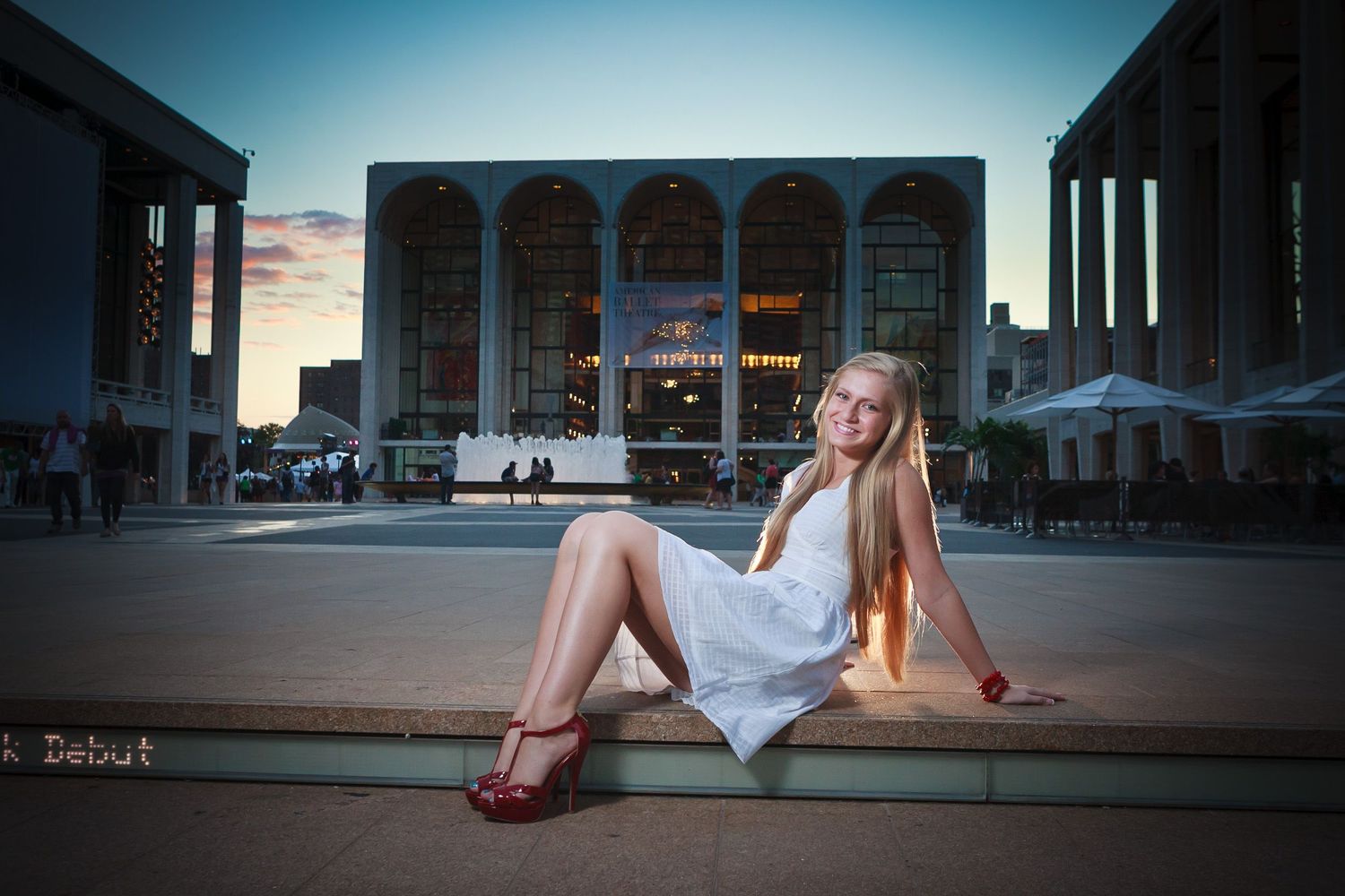 Blonde teenage girl in white dress sitting at Lincoln Center plaza at dusk.