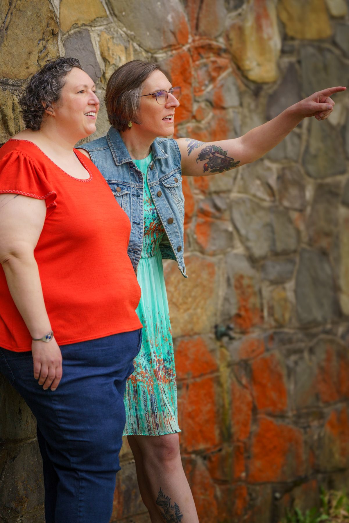 Two women stand by a stone wall, one in red top pointing into the distance, both smiling outdoors.