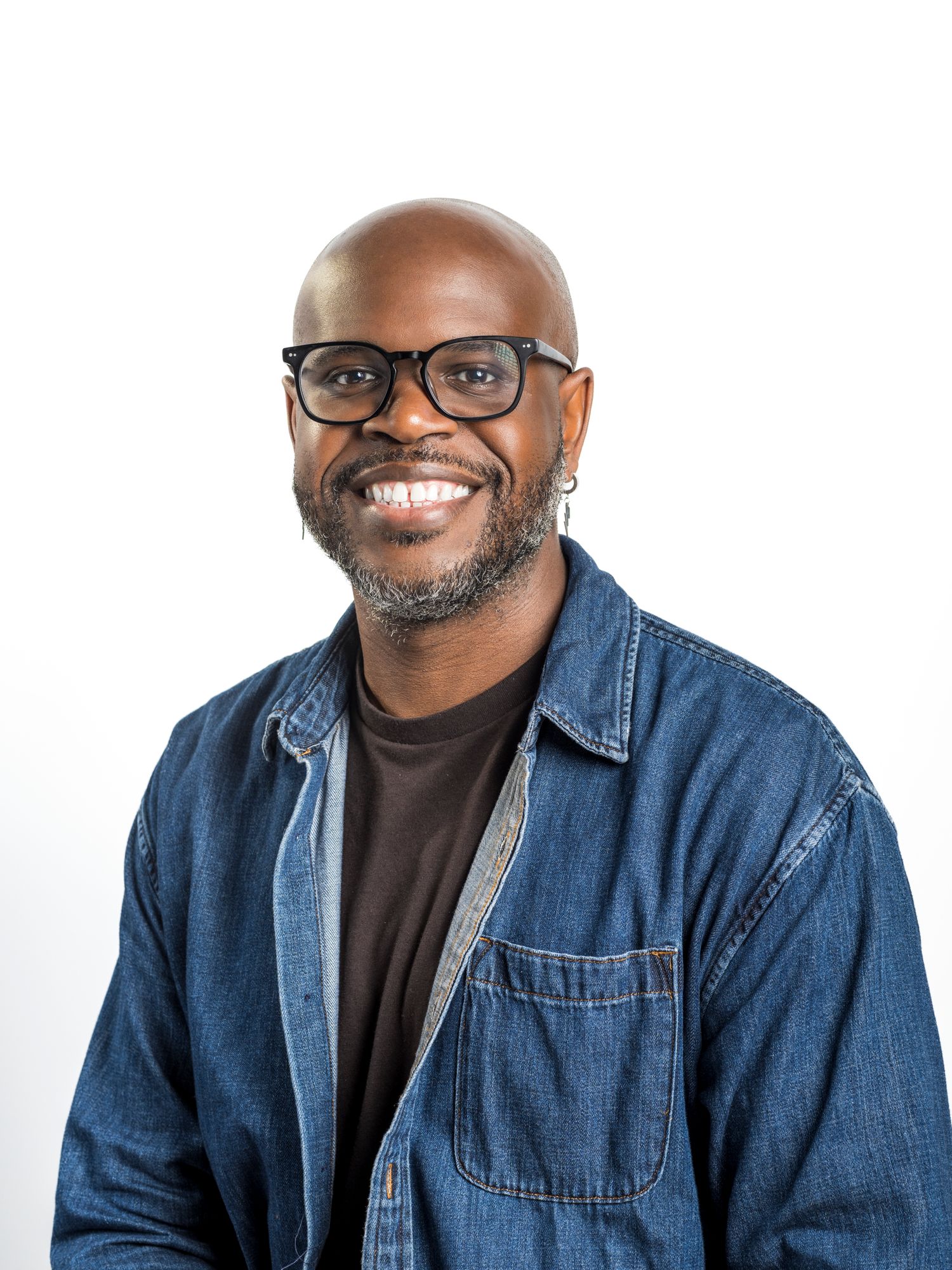 Smiling bald Black man with glasses and beard wearing a denim jacket over a dark shirt against white background.
