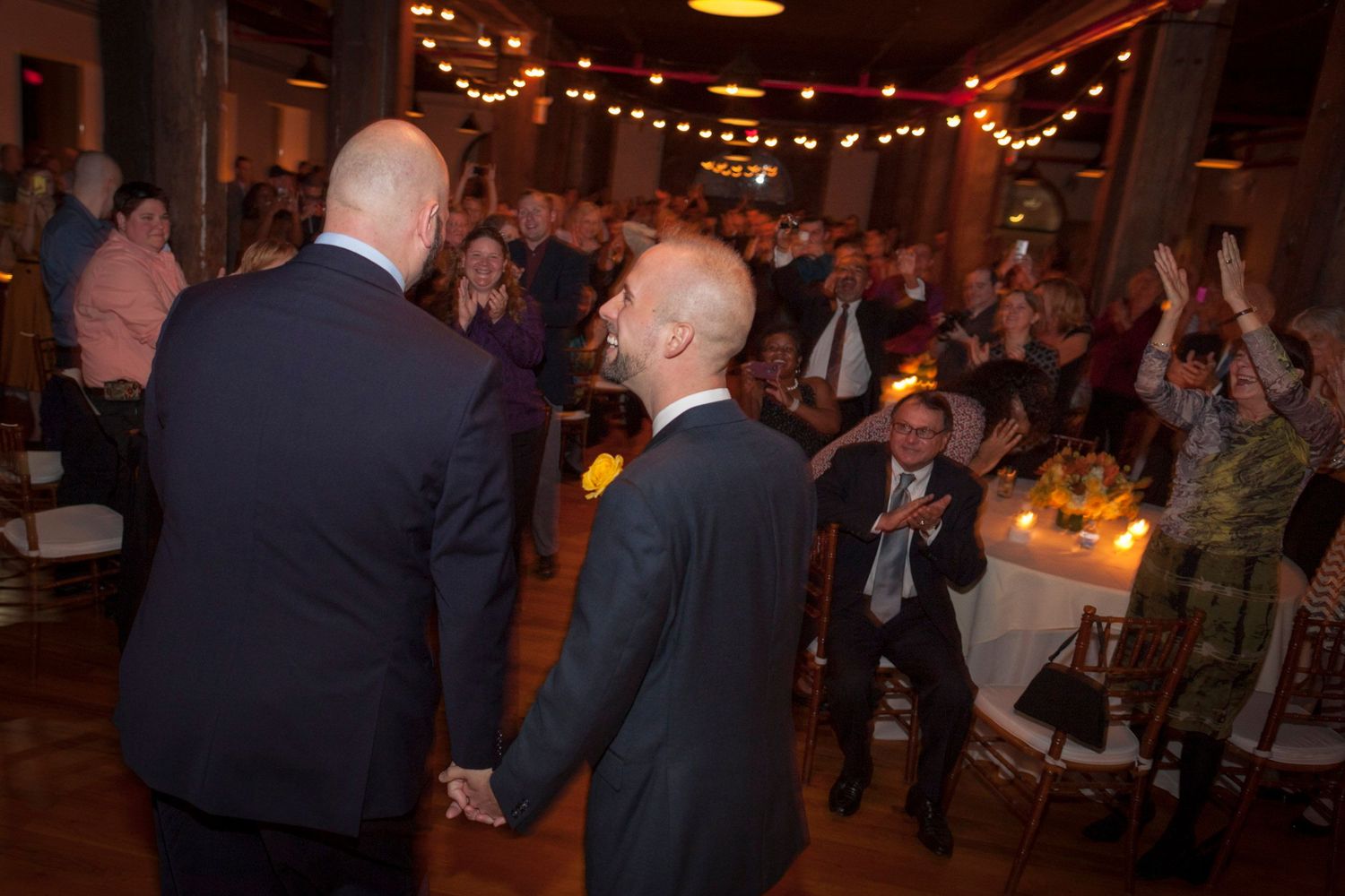 Two grooms holding hands as wedding guests applaud in warmly lit reception venue.
