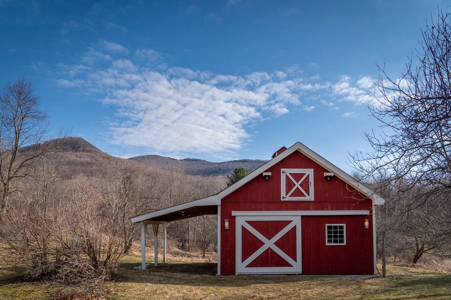 Red barn with white trim and X-pattern doors set against blue sky and mountain landscape in rural countryside.
