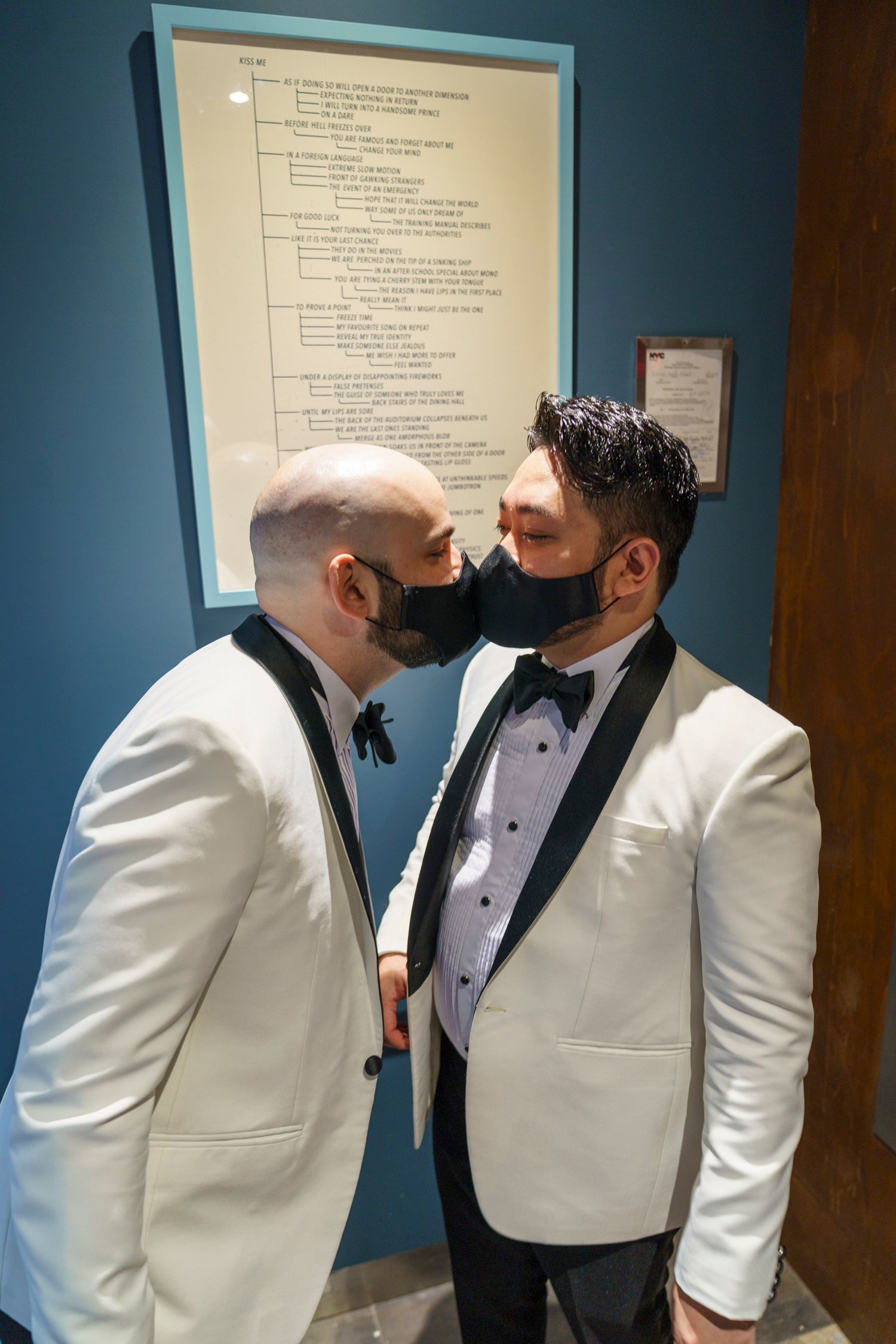 Two grooms in white tuxedos share a masked kiss at their wedding ceremony.