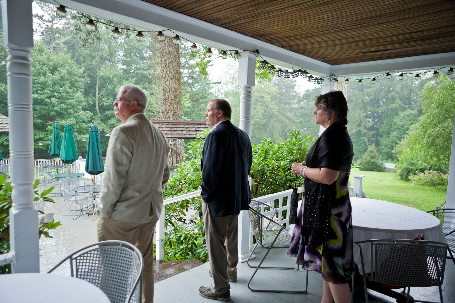 Three guests standing on a covered porch overlooking a lush green garden at an outdoor event.