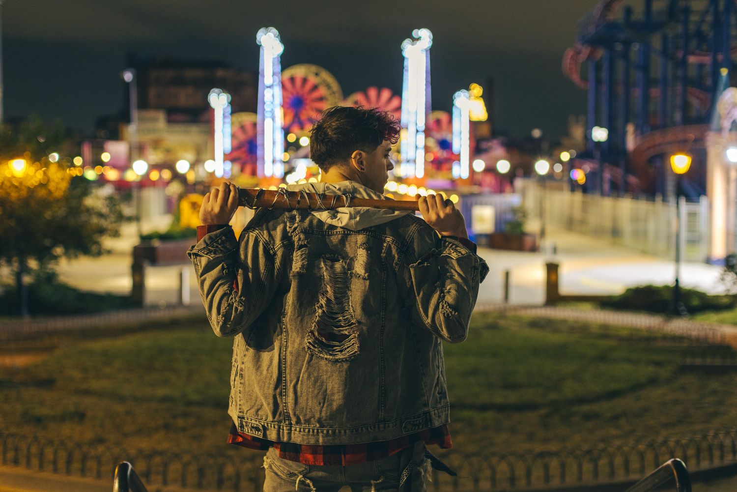 Man in an olive jacket stands with his back to the camera, holding a stick across his shoulders, facing a lit amusement park at dusk.